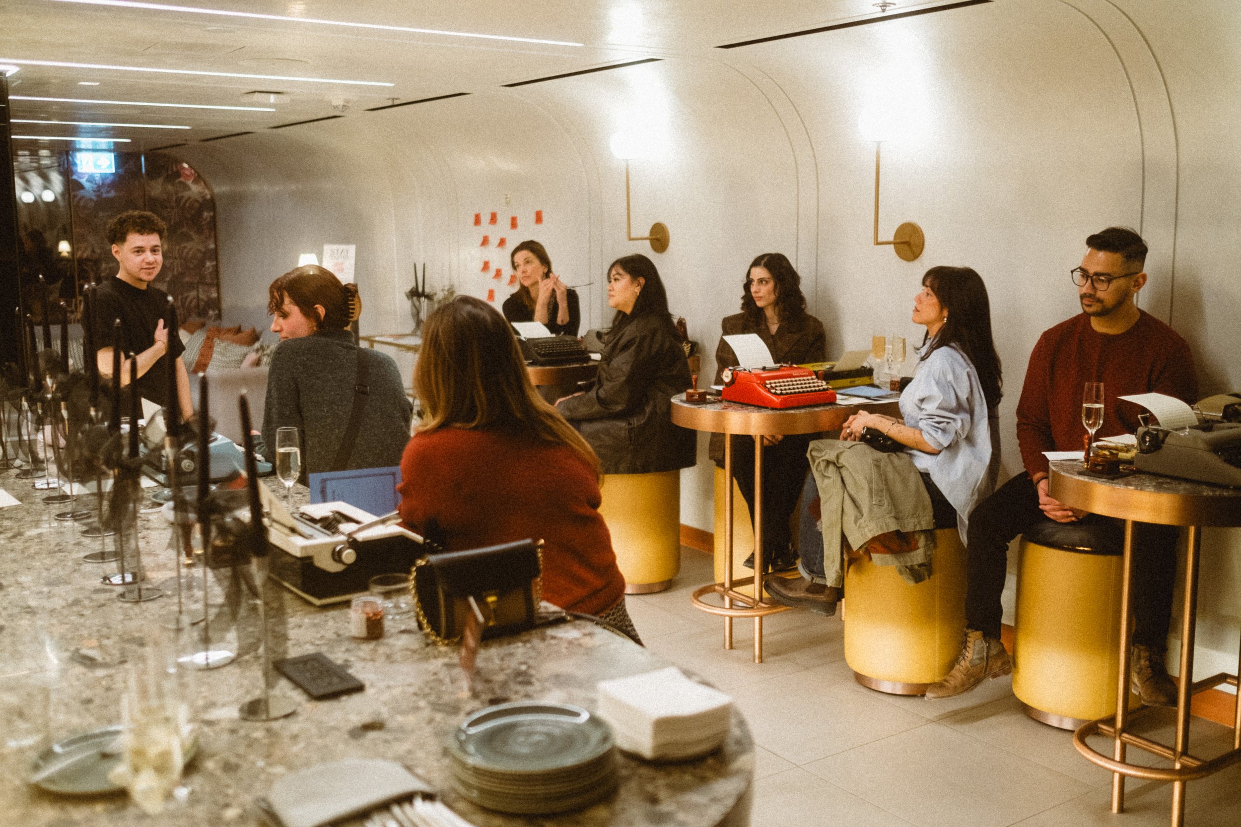 A group of people sit around the bar in a dimly lit restaurant or cafe, listening to a person speak or perform. The setting has a modern, cozy ambiance with warm lighting and decorative wall accents.