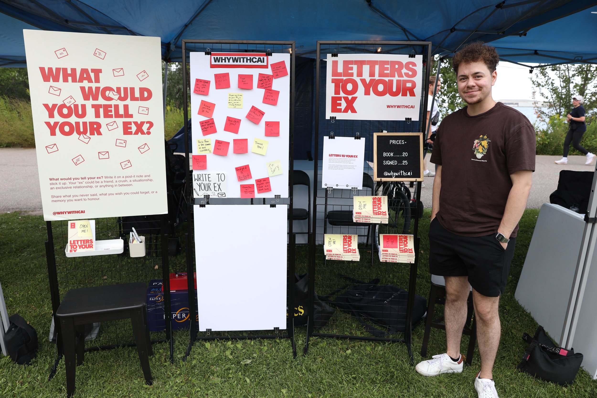 A man standing outdoors under a large canopy, smiling at the camera, near a display booth with signs and notes about writing letters to an ex, including a large sign with red text asking, 'What would you tell your ex?' and various handwritten notes attached to a whiteboard.