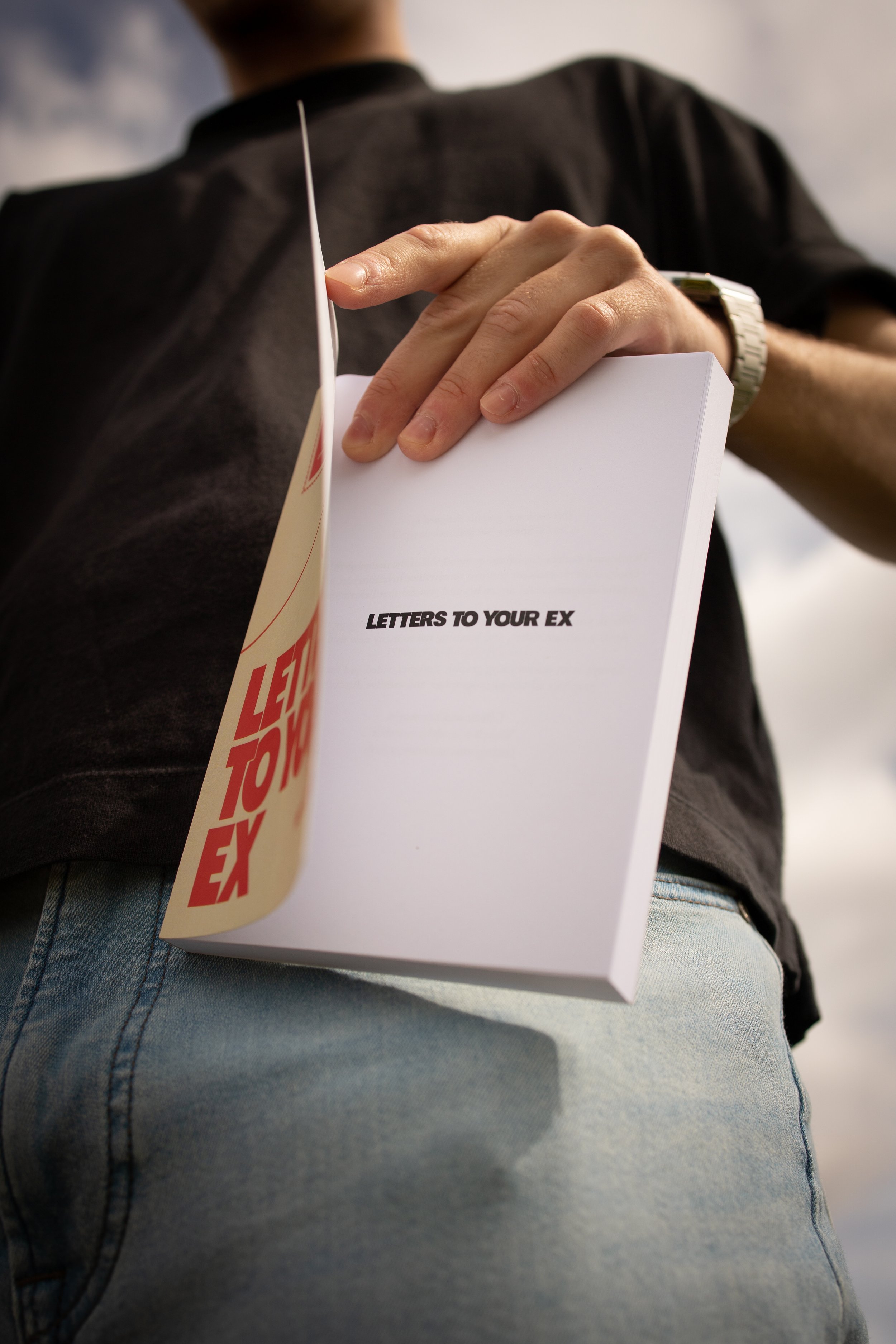 A person wearing a black shirt and light blue jeans is holding a white notepad or booklet titled 'LETTERS TO YOUR EX' in their right hand, with the person's back facing the camera.