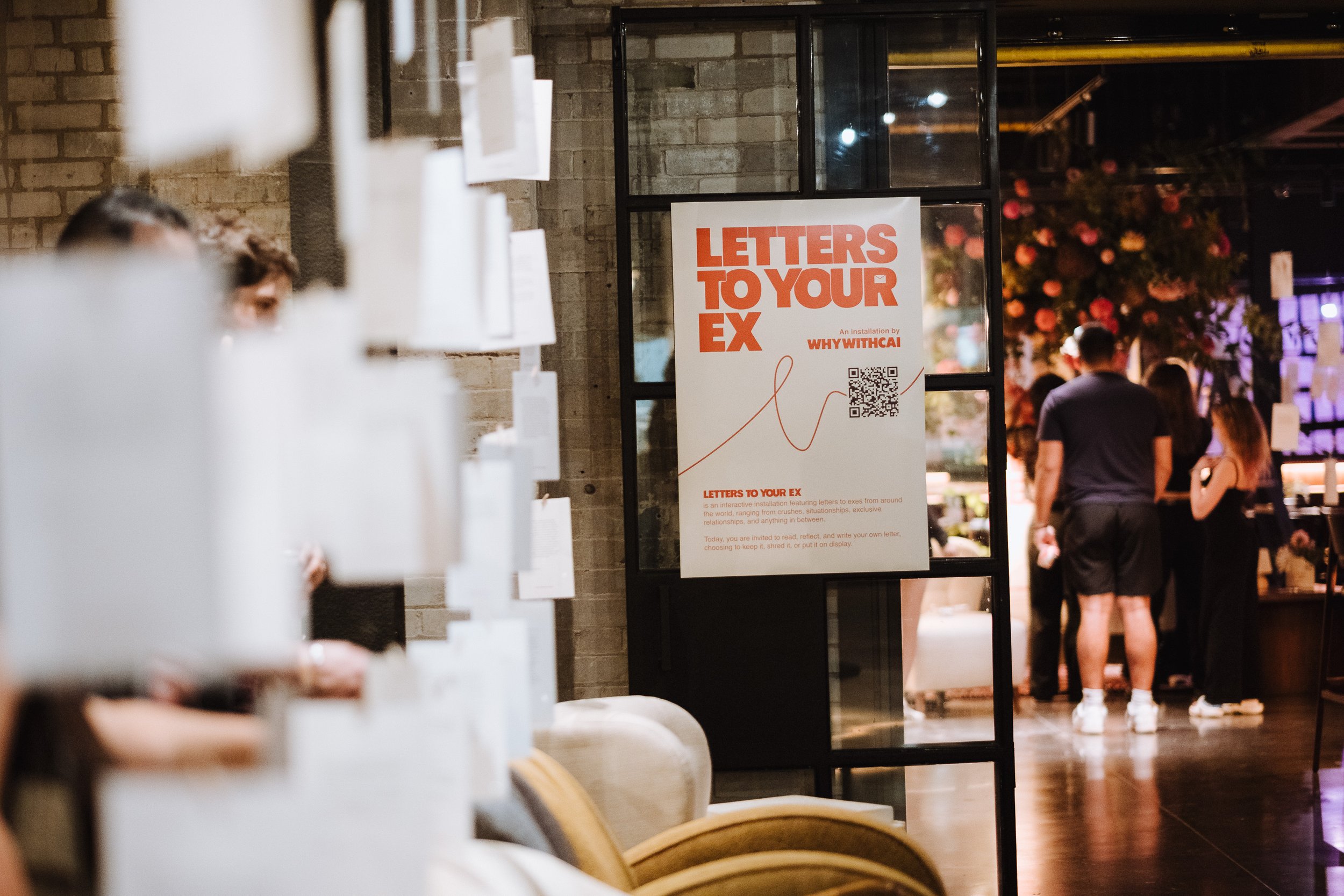 Sign on a window advertising an exhibition called "Letters to Your Ex" by WHYWITHCAI, with a QR code below the text. In the background, there are people walking outside in an urban setting at night, with some flower arrangements visible.