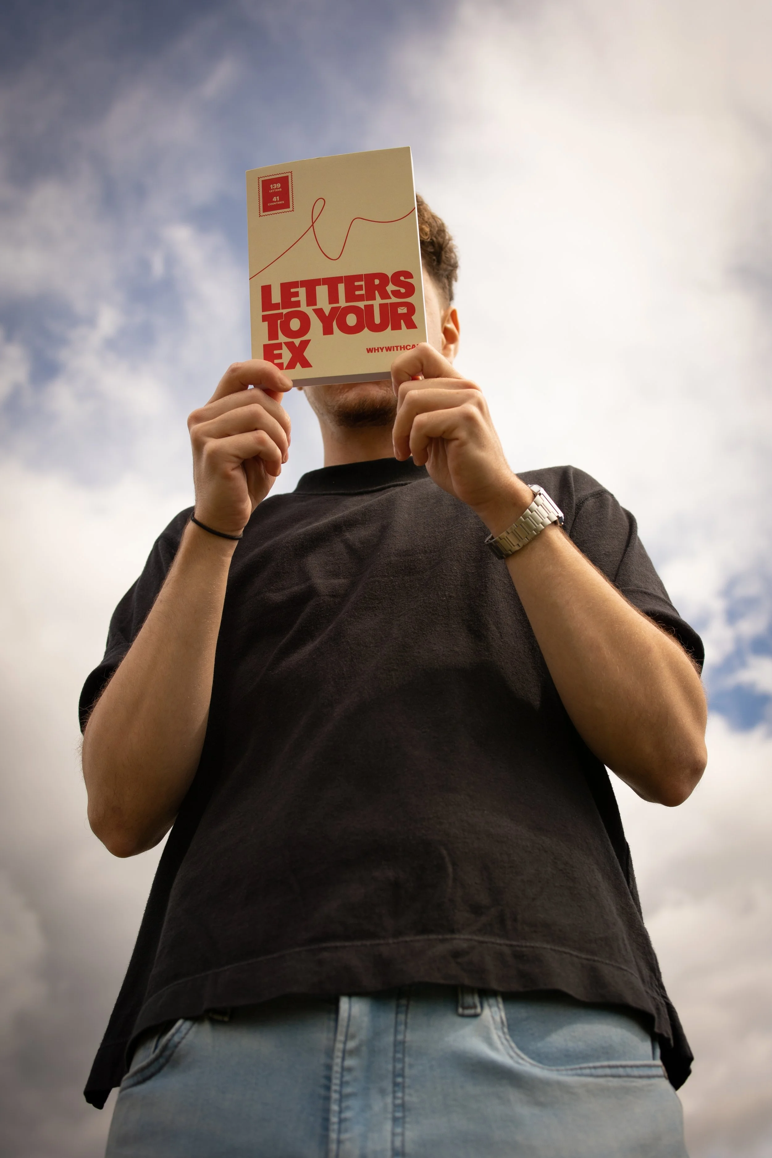 Person holding a booklet titled 'Letters to Your Ex' covering their face, wearing a black shirt and light blue jeans, against a cloudy sky background.