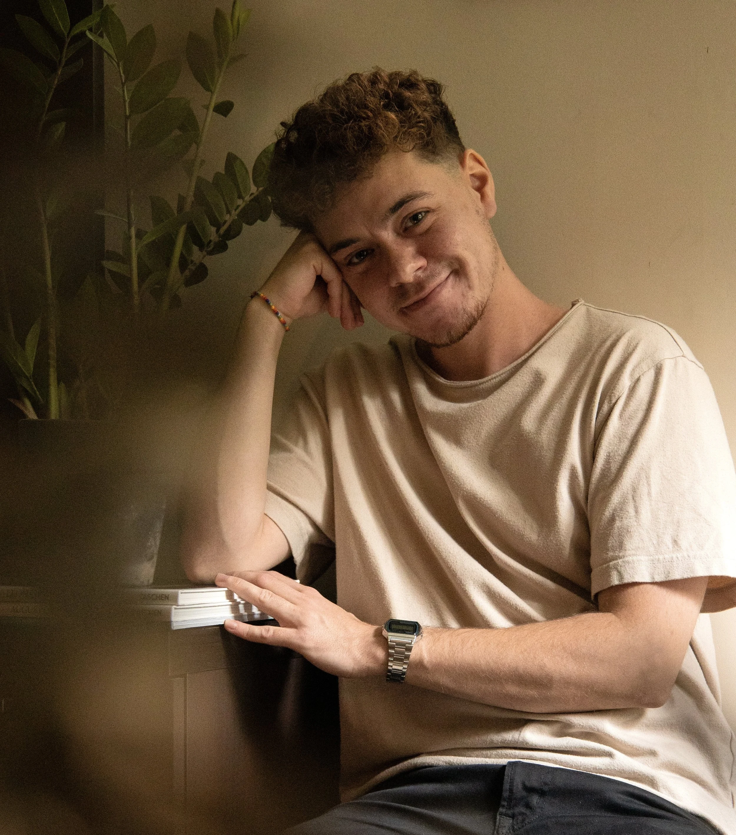 A young man with curly hair is sitting at a table, resting his head on his hand, smiling softly at the camera. He is wearing a beige T-shirt, a silver wristwatch, and a colorful beaded bracelet. There is a potted plant with green leaves on the table to his left, and the background is a plain, light-colored wall.