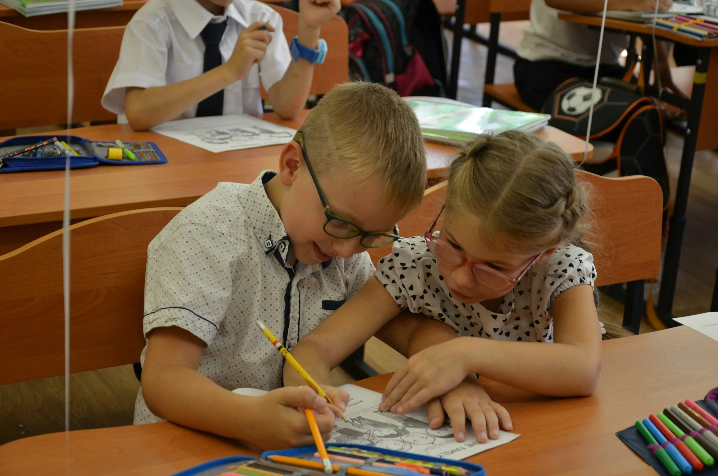 Two young children, a boy and a girl, are sitting at a desk working on coloring books. They are sharing a set of colored pencils and seem to be focused on their activity. In the background, other children are visible, some with backpacks and school supplies, indicating a classroom setting.