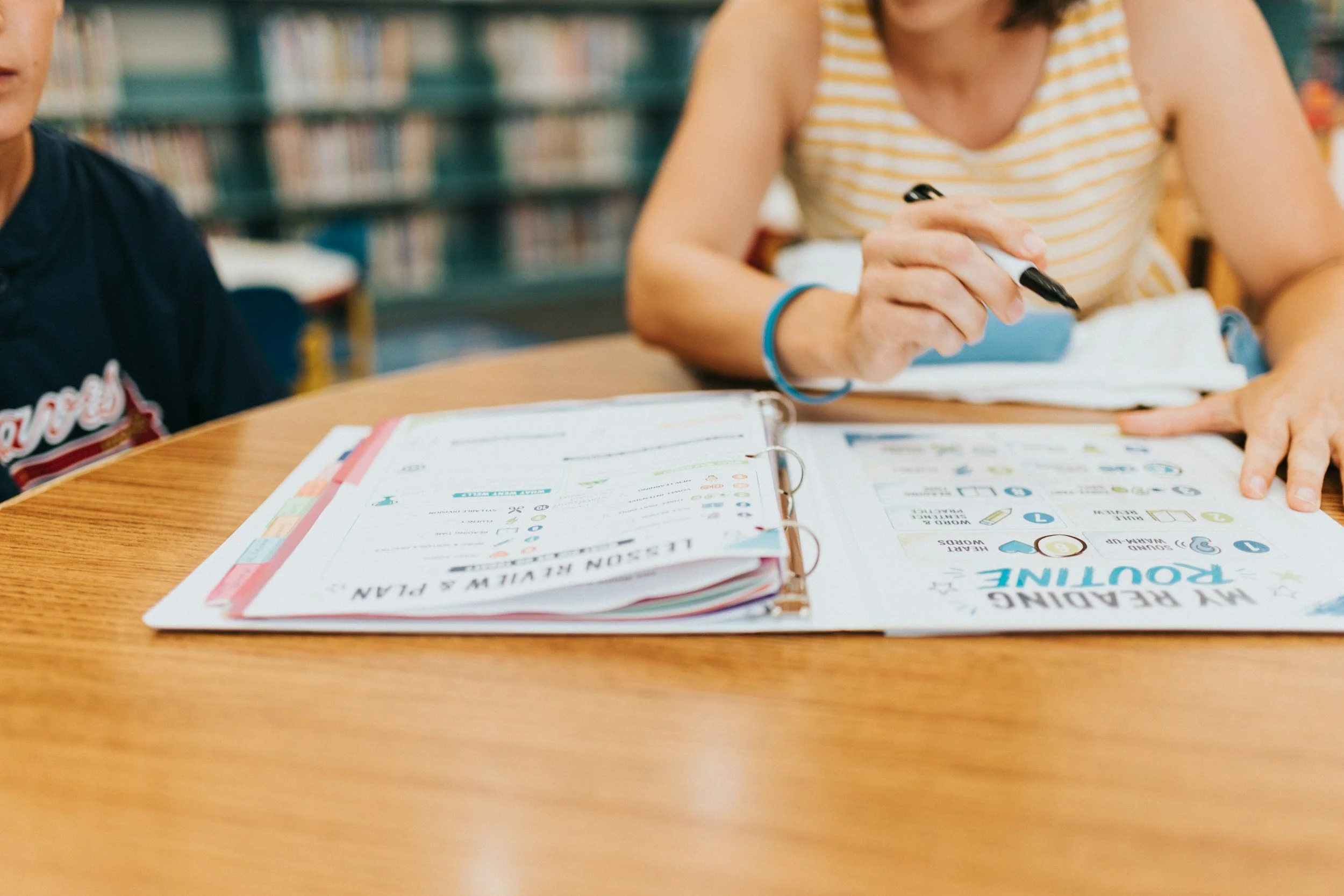 Close-up of a person writing with a black marker in a workbook on a wooden table, with educational materials and a second person partially visible in a library or classroom setting.