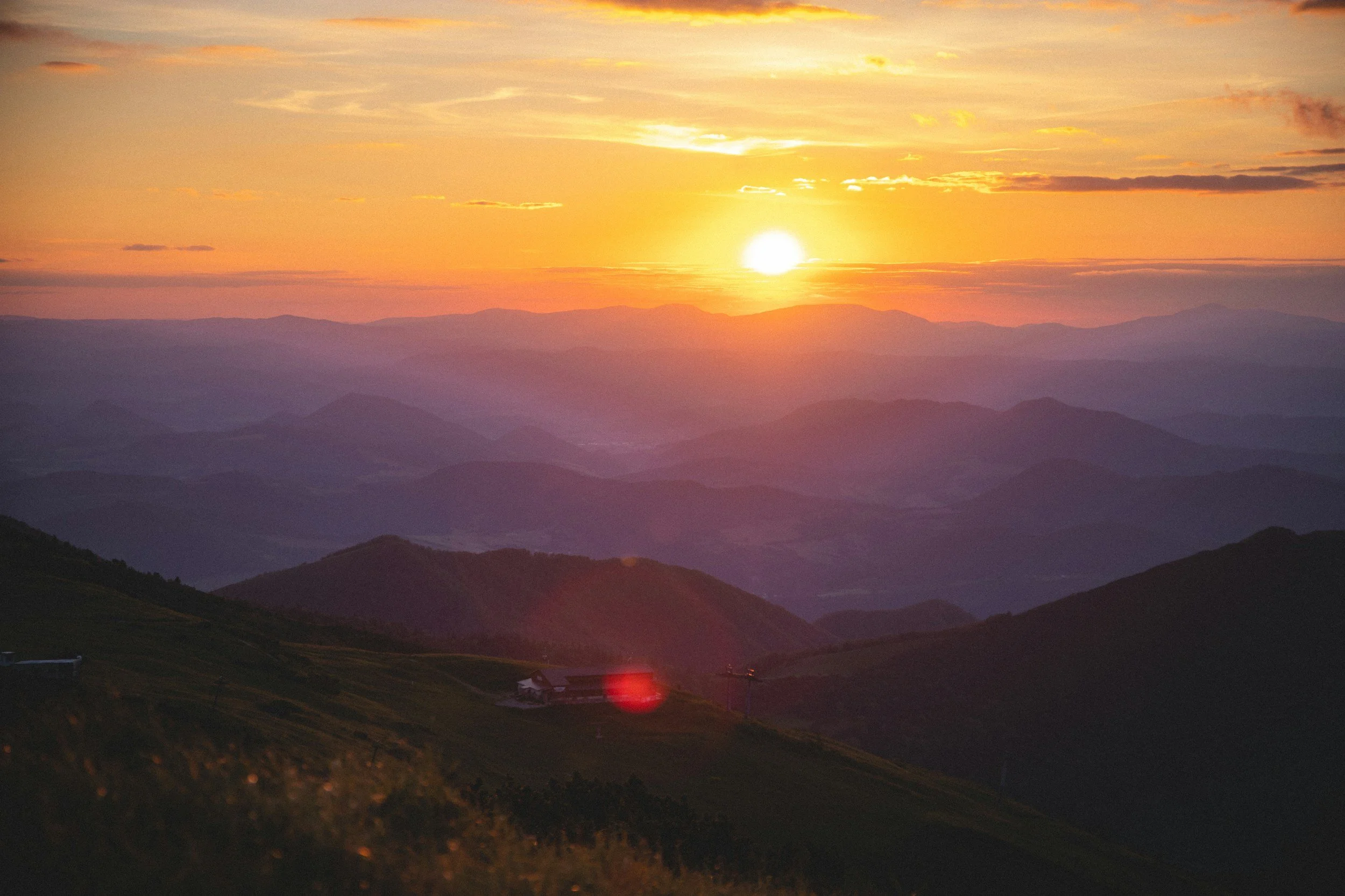 A scenic view of a sunset over mountains with colorful sky and a few clouds, with dark rolling hills in the foreground.