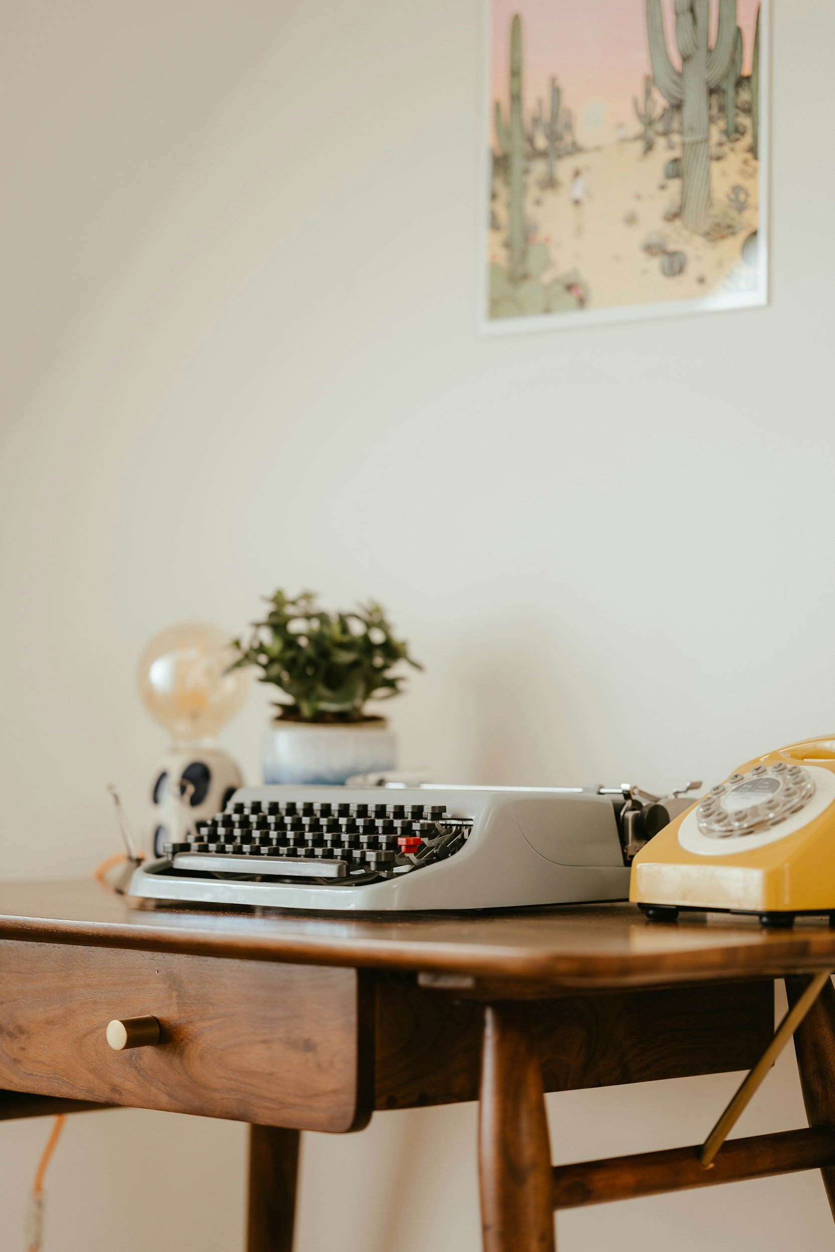 A vintage yellow rotary telephone on a wooden desk with a typewriter, plant, and wall decor in the background.