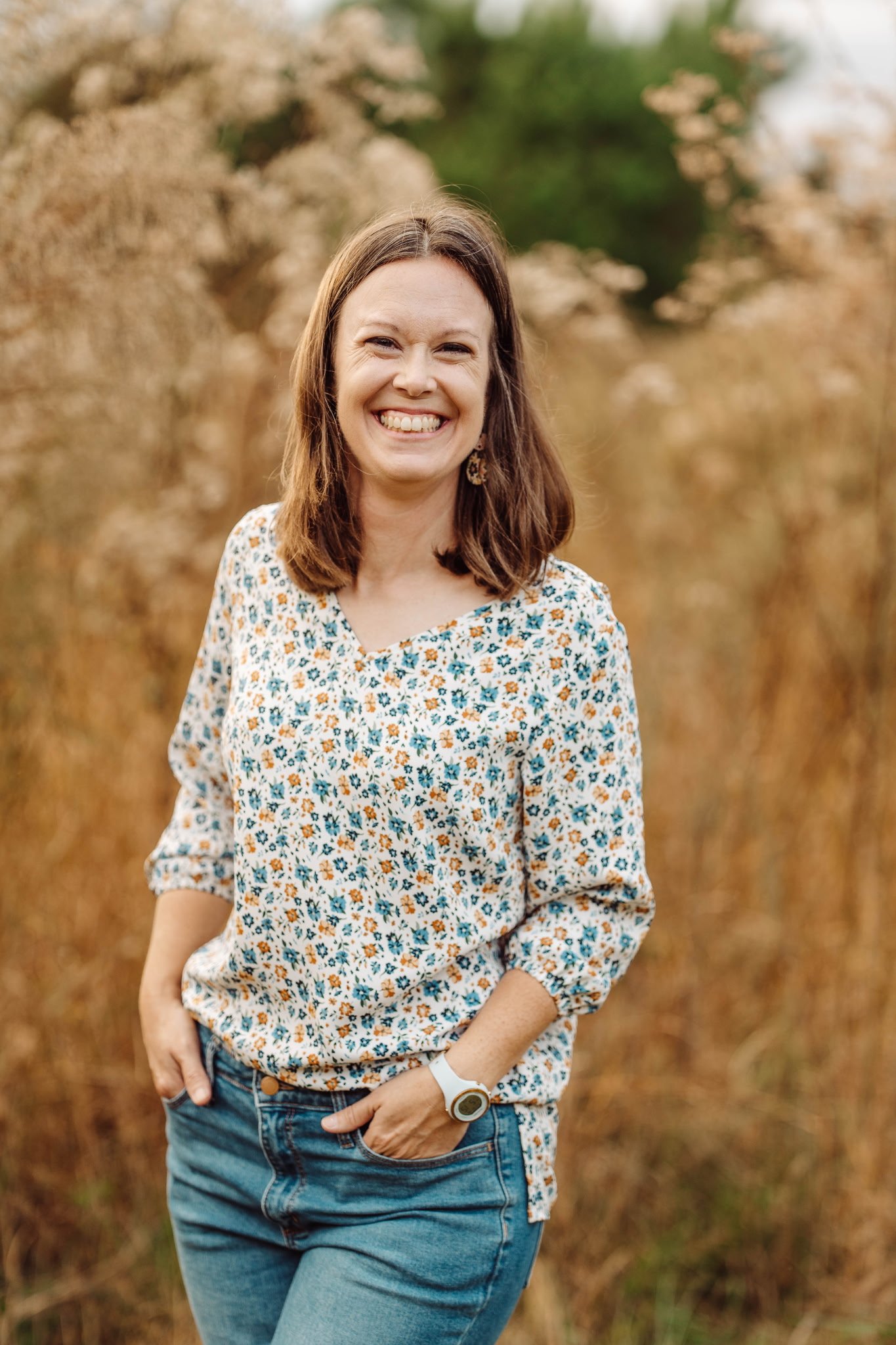 A smiling woman with shoulder-length brown hair stands outdoors in a field with tall, golden brown plants and green trees in the background. She is wearing a floral-patterned blouse, blue jeans, a white watch, and earrings.