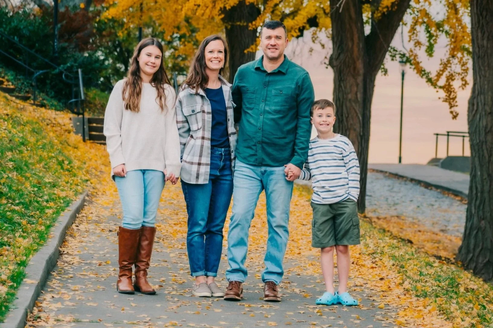 Family of five walking on a leaf-covered sidewalk in autumn, surrounded by trees with yellow and orange leaves, holding hands and smiling.