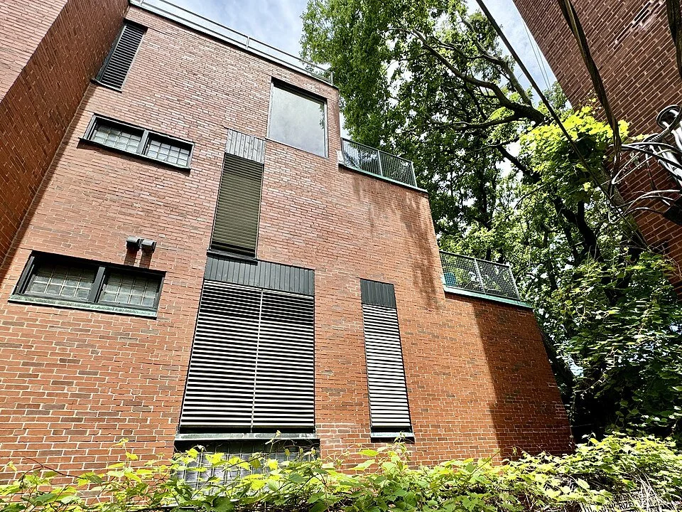 Low-angle view of a multi-story brick building with various windows and metal shutters, surrounded by green trees and foliage.