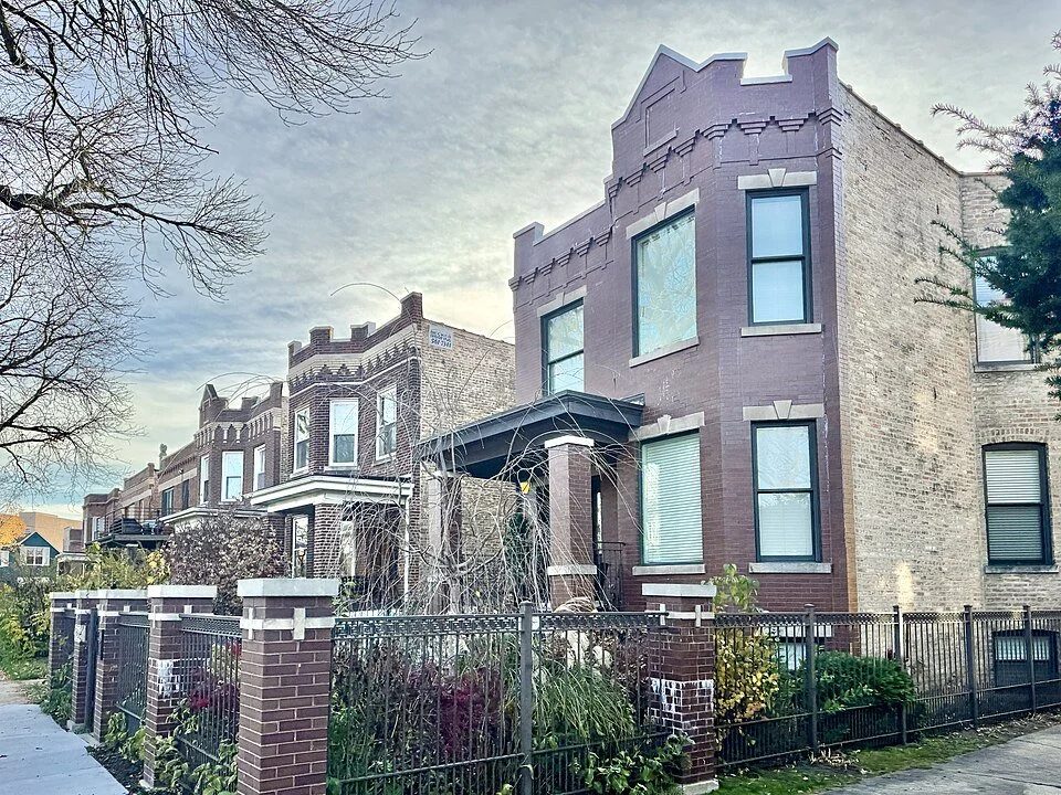 A row of historic brownstone buildings with decorative brickwork, large windows, and iron fences, set on a city street during daytime with overcast sky.