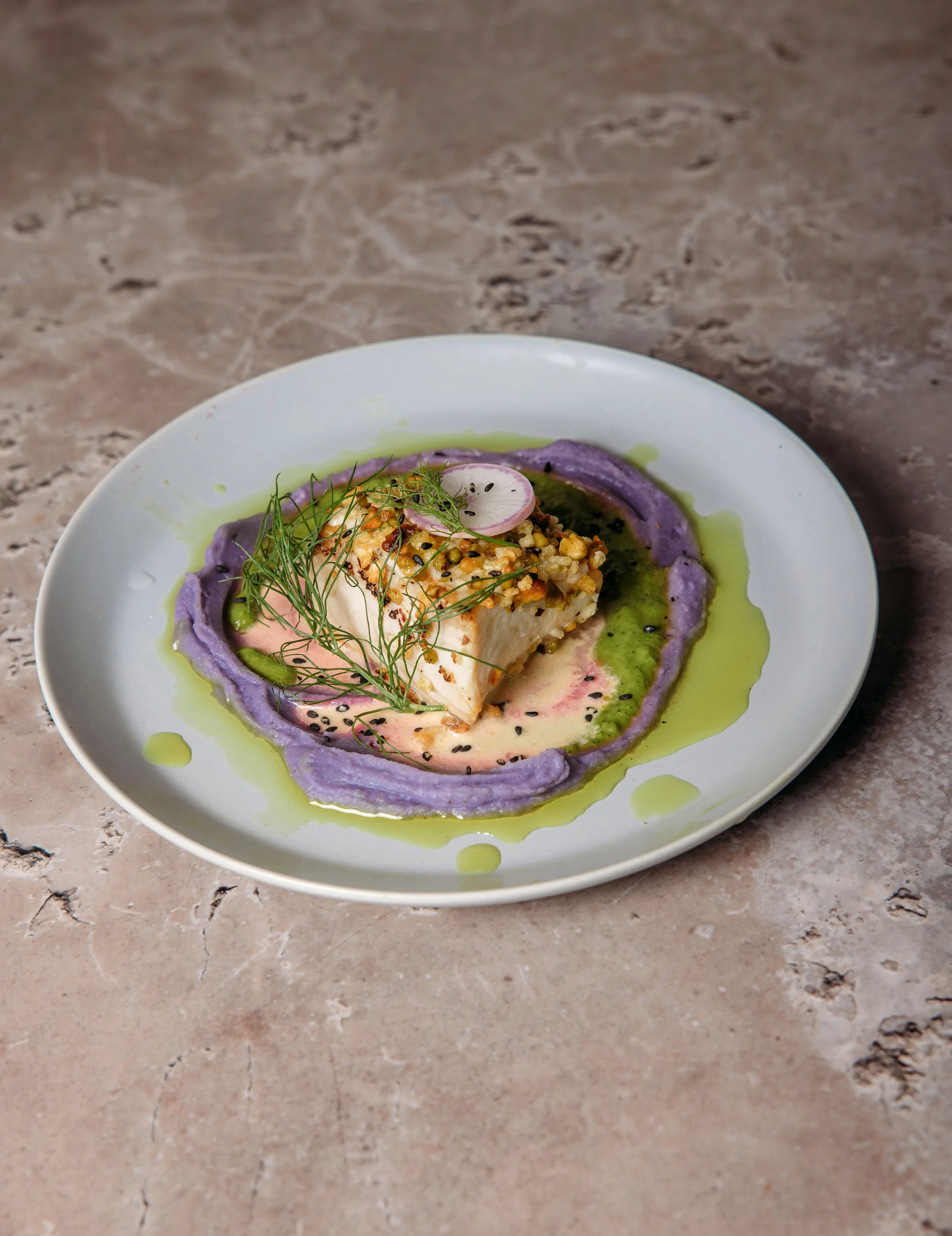 A plated dish featuring a piece of white fish topped with herbs and seeds, surrounded by purple and green sauces, garnished with radish slices and herbs on a white plate.