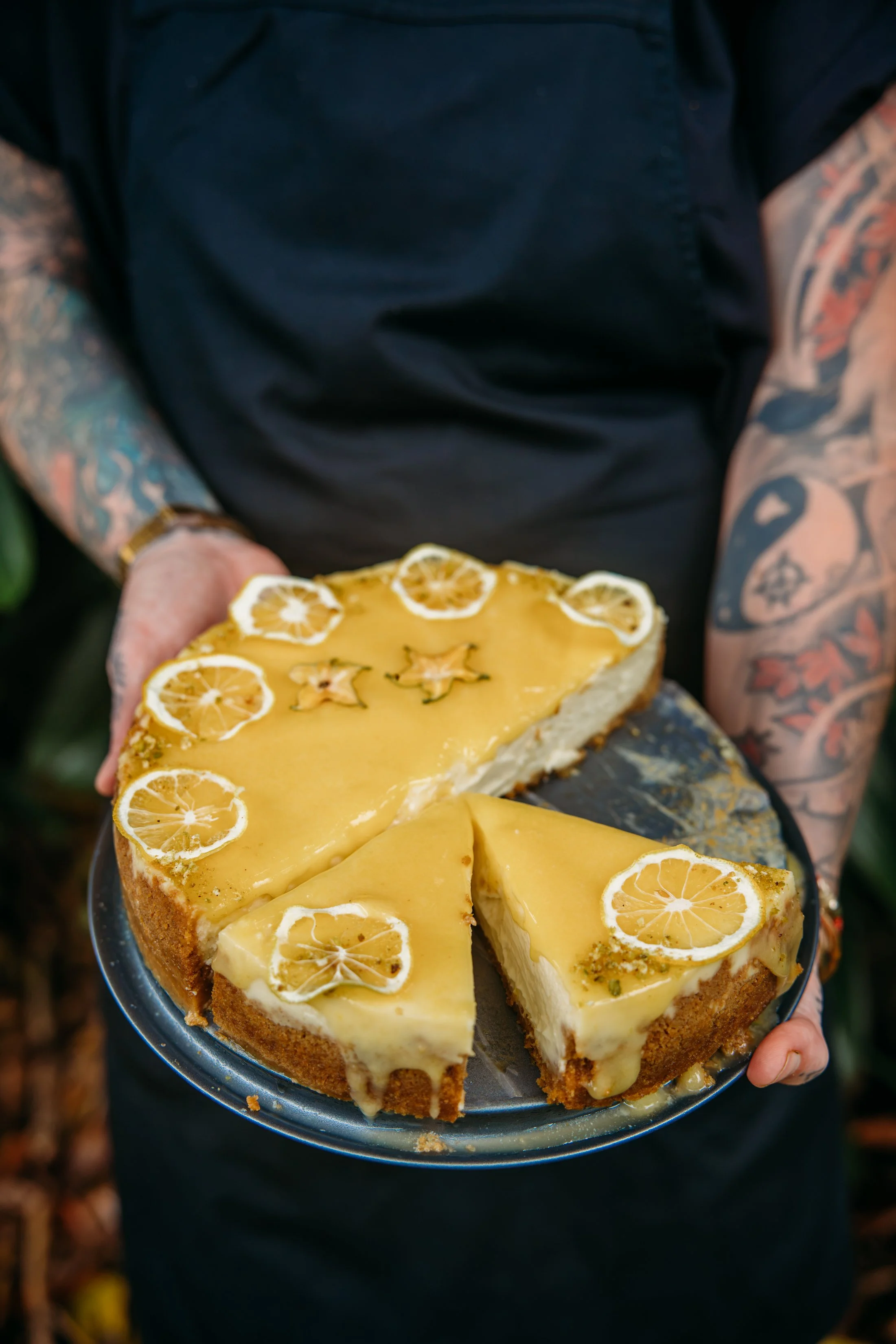 A person with colorful tattoos on both arms holds a tray with a lemon-flavored cake topped with lemon slices.
