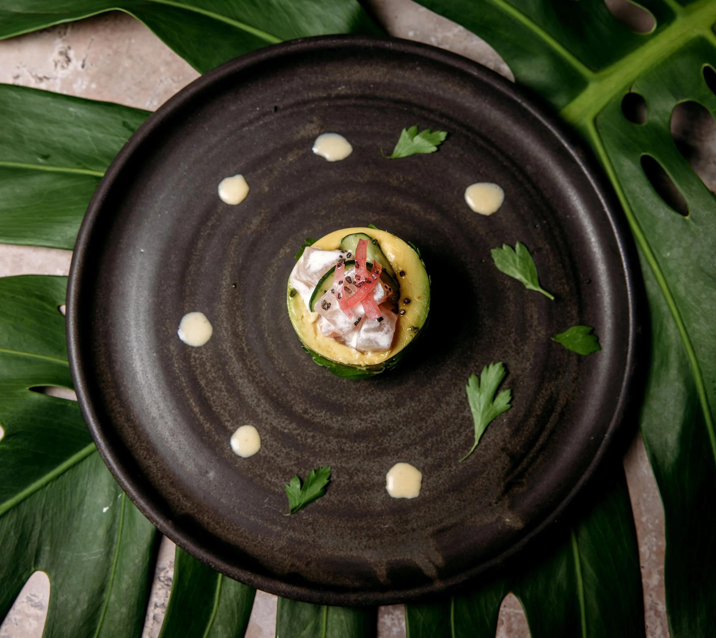 A dark round plate with a gourmet appetizer topped with cucumber slices, red pepper strips, and herbs, garnished with small white sauce dots and surrounded by large green leaves on a textured surface.