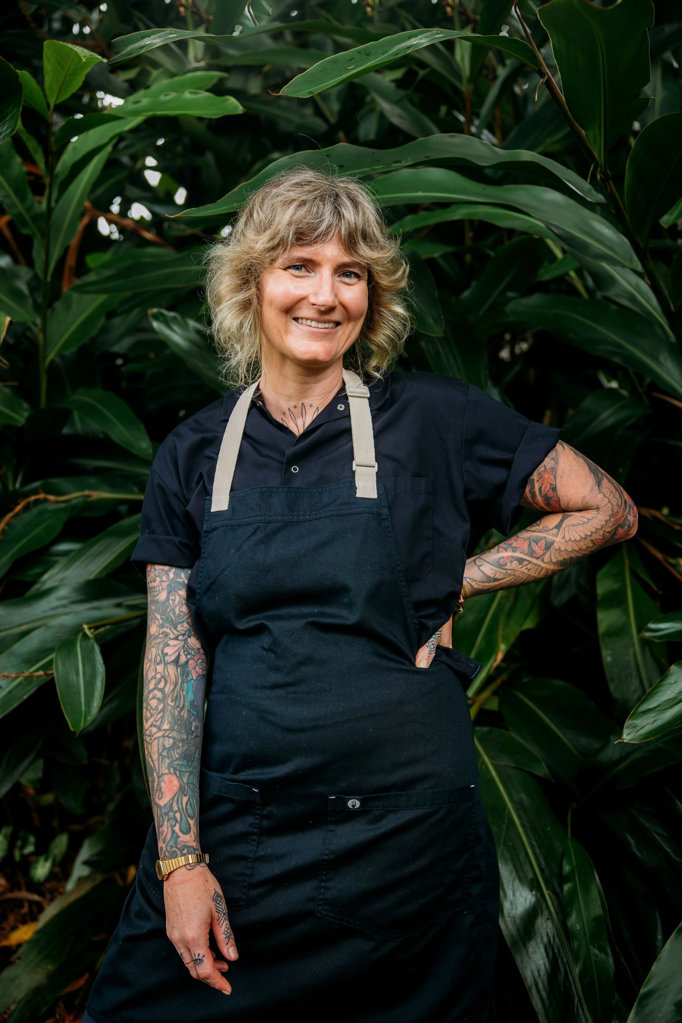 A woman with blonde curly hair and tattoos on her arms, wearing a black shirt and apron, standing in front of large green leaves.