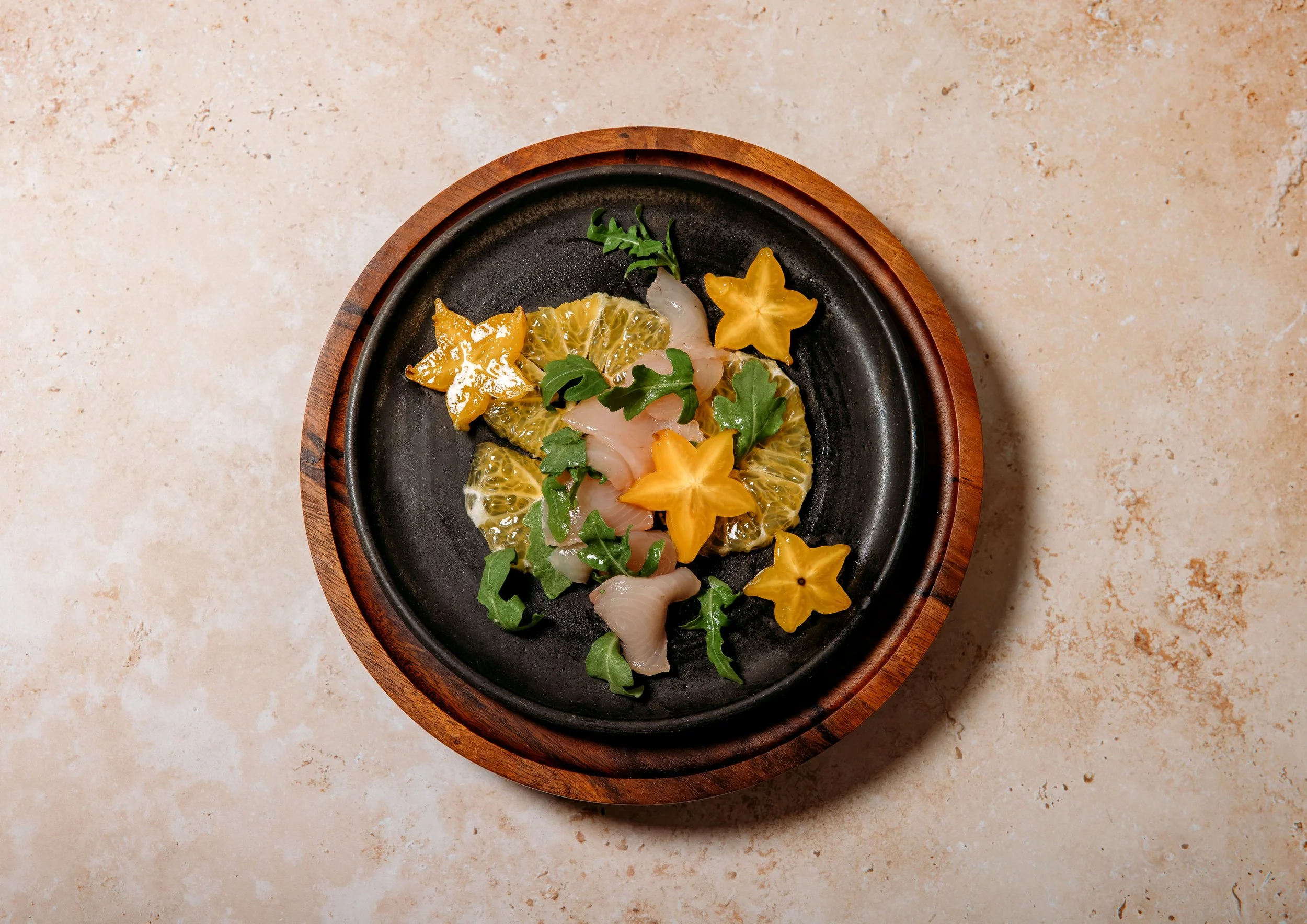 A black plate with a wooden rim containing thinly sliced transparent fish, yellow star-shaped fruit slices, green arugula leaves, and slices of yellow citrus fruit, placed on a beige textured surface.