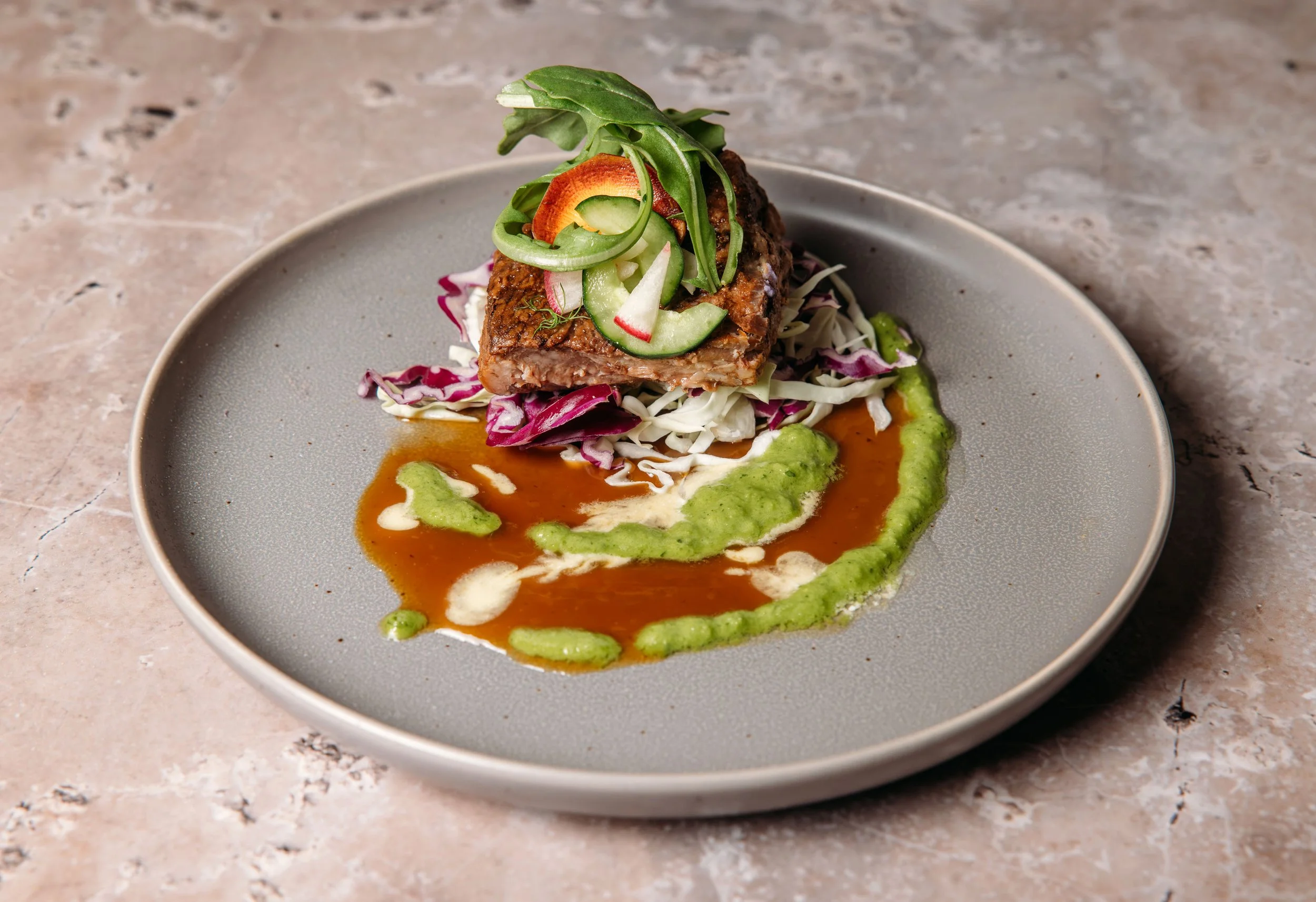 A plated dish featuring a piece of grilled meat topped with sliced cucumbers, radishes, and greens, served with colorful sauces and shredded cabbage on a gray ceramic plate.