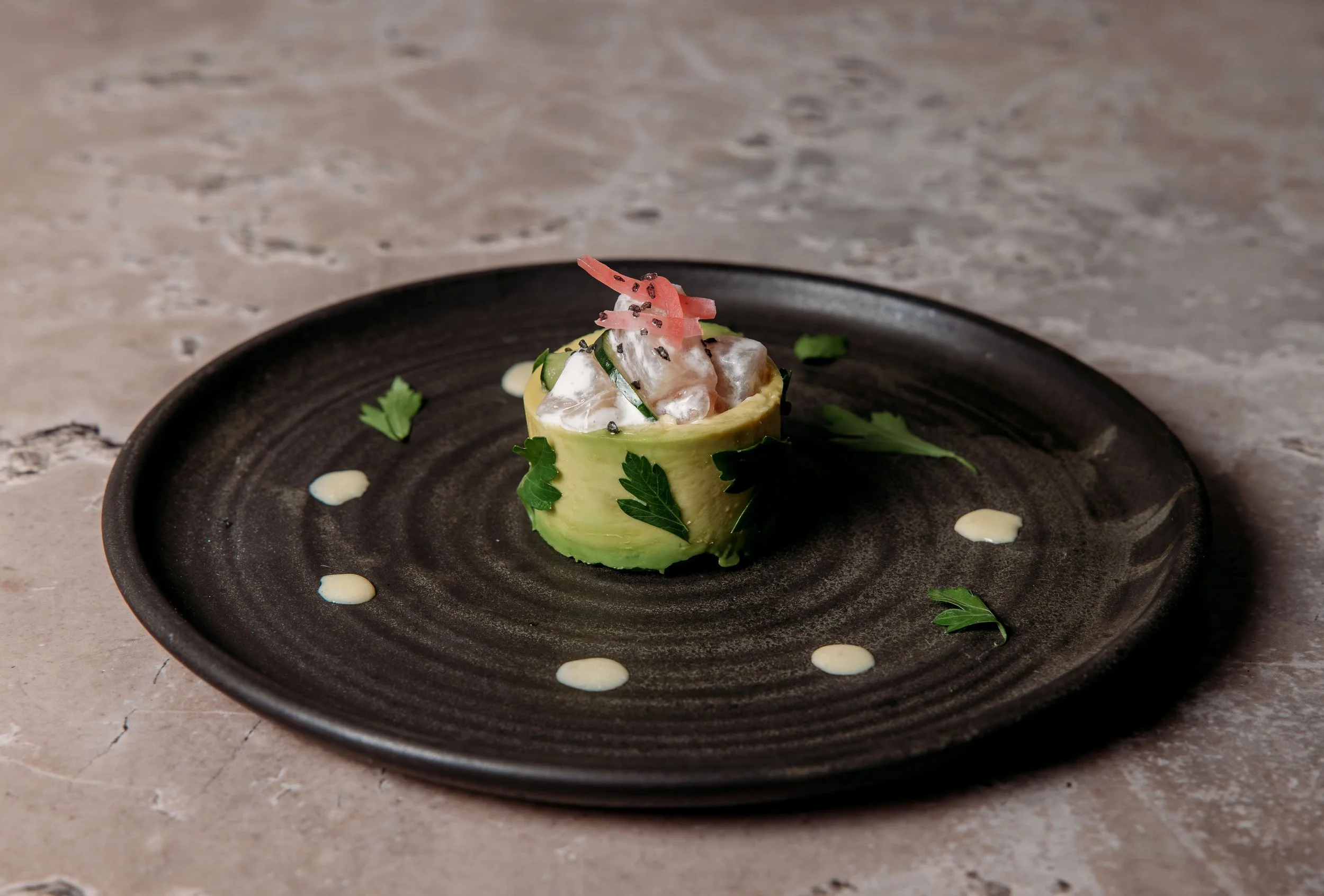 A small savory tart garnished with pink pickled ginger, black sesame seeds, and green herbs on a black plate.