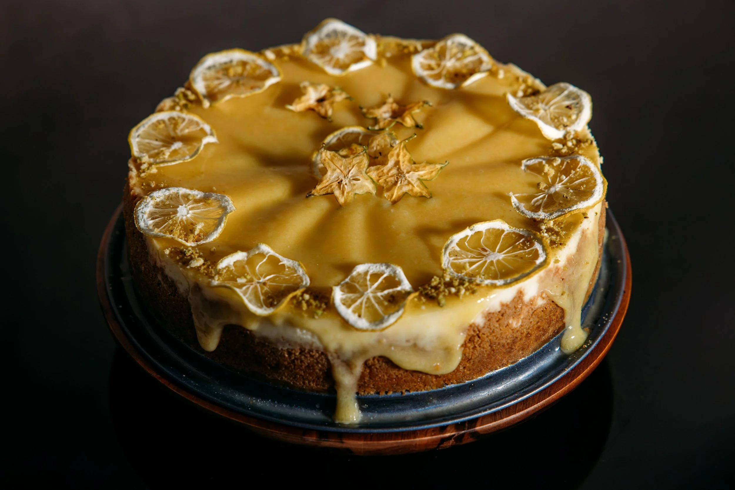 A round cake topped with a smooth yellow glaze, decorated with dried lemon slices around the edges and star-shaped decorations in the center, sitting on a dark surface.