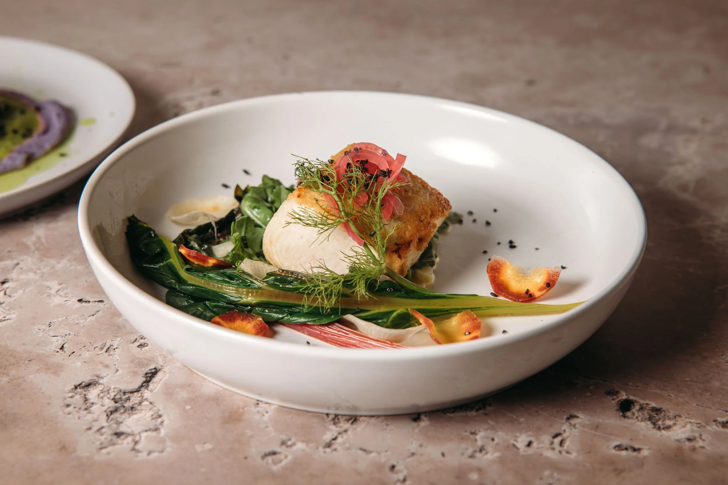 Dish featuring grilled fish on a bed of greens garnished with herbs, sliced pickled onions, and pepper flakes, served in a white bowl.