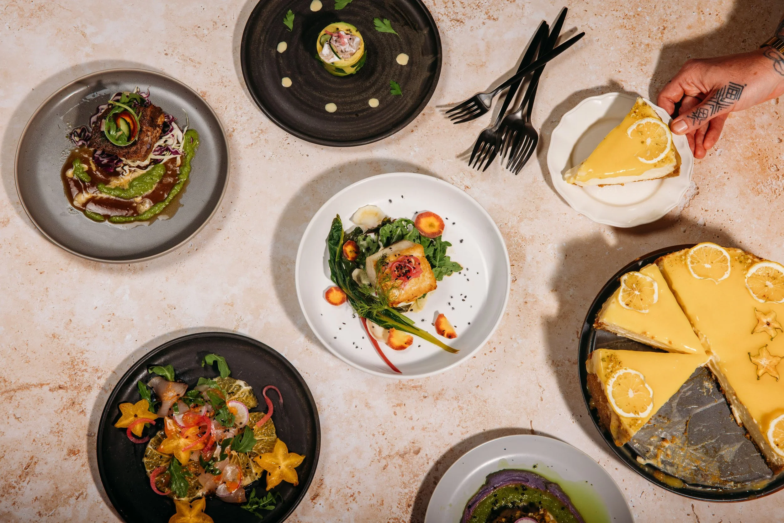 Various plates of food on a light-colored table, including a slice of lemon cake, a vegetable salad, a fish dish, and small appetizer portions.