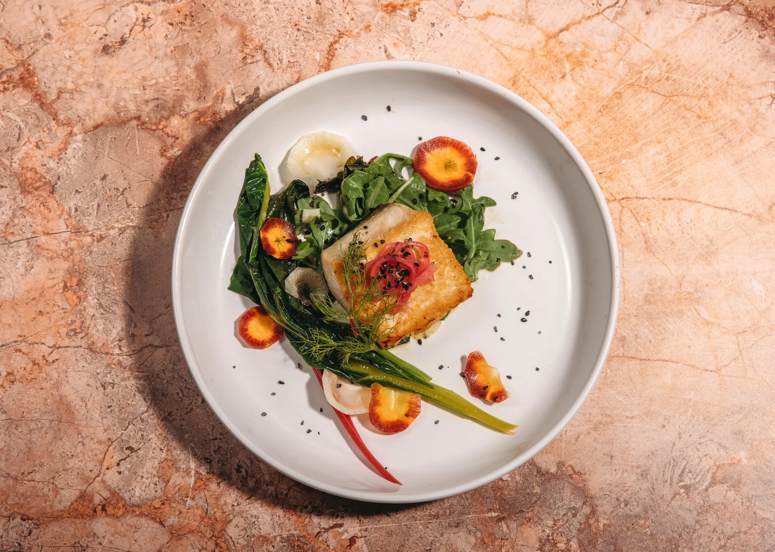 A white oval plate with a portion of grilled fish topped with sliced tomatoes, garnished with herbs and black sesame seeds, resting on a pinkish-orange textured surface.