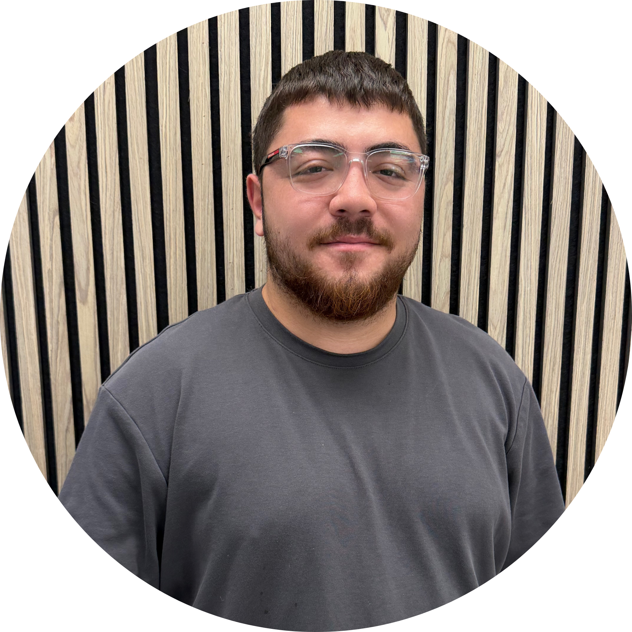 Close-up of a man with short dark hair, glasses, and a beard, smiling, against a background of vertically striped light wood and black panels.