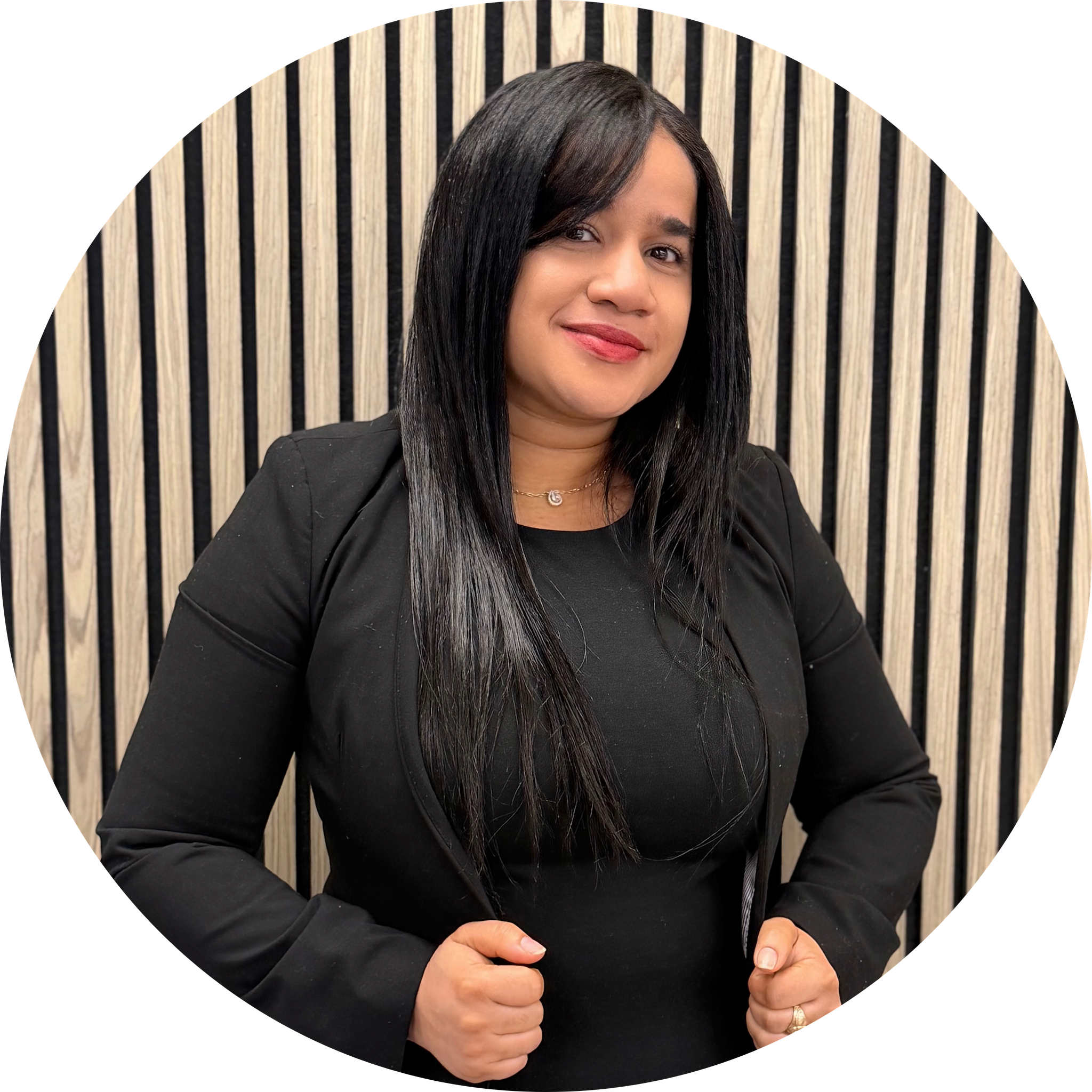 A woman with long black hair wearing a black blazer smiling and giving a thumbs-up gesture in front of a wooden slat background.