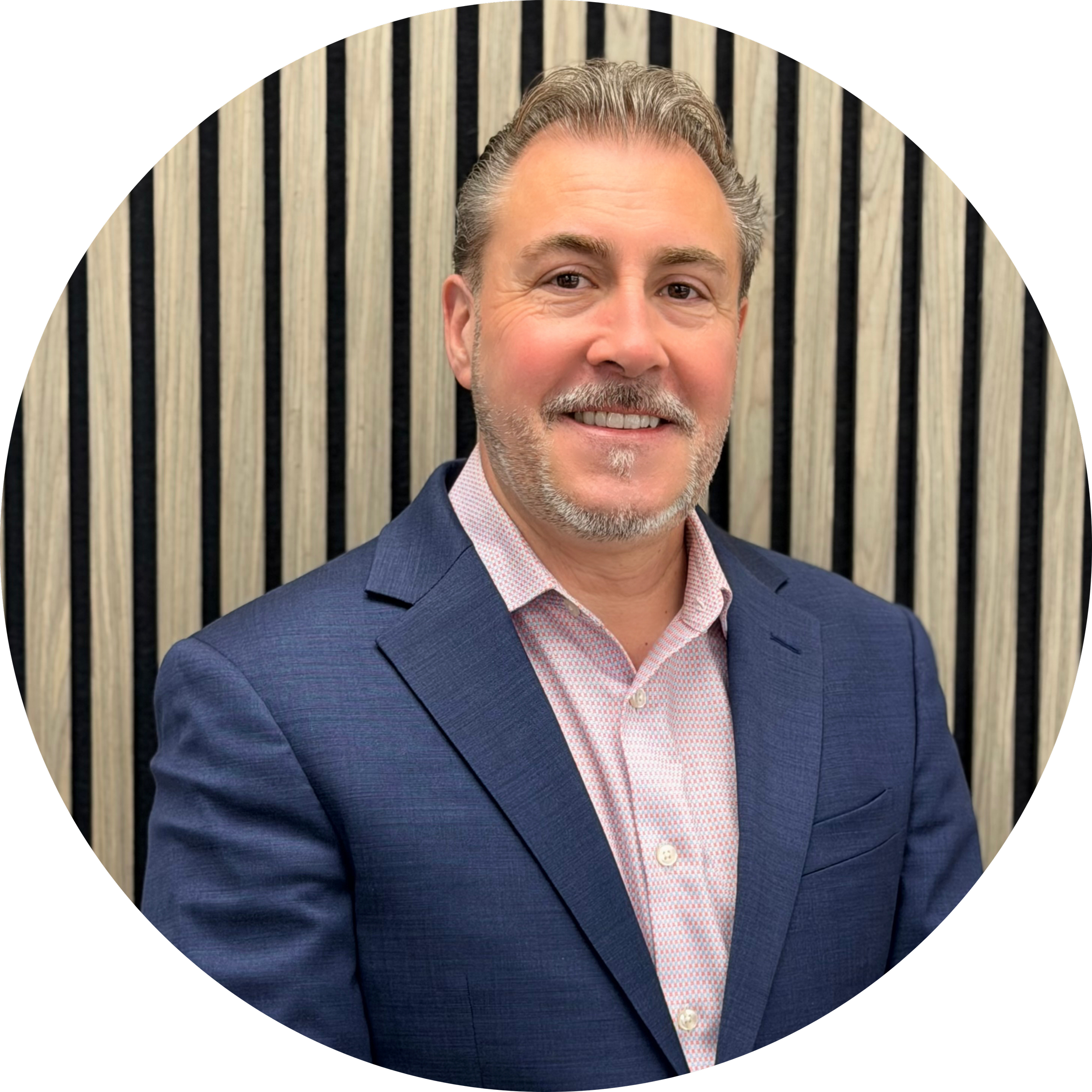 Portrait of a middle-aged man with gray hair and a beard, smiling, wearing a dark blue suit and a pink and white checkered shirt, standing in front of a background with vertical black and beige stripes.