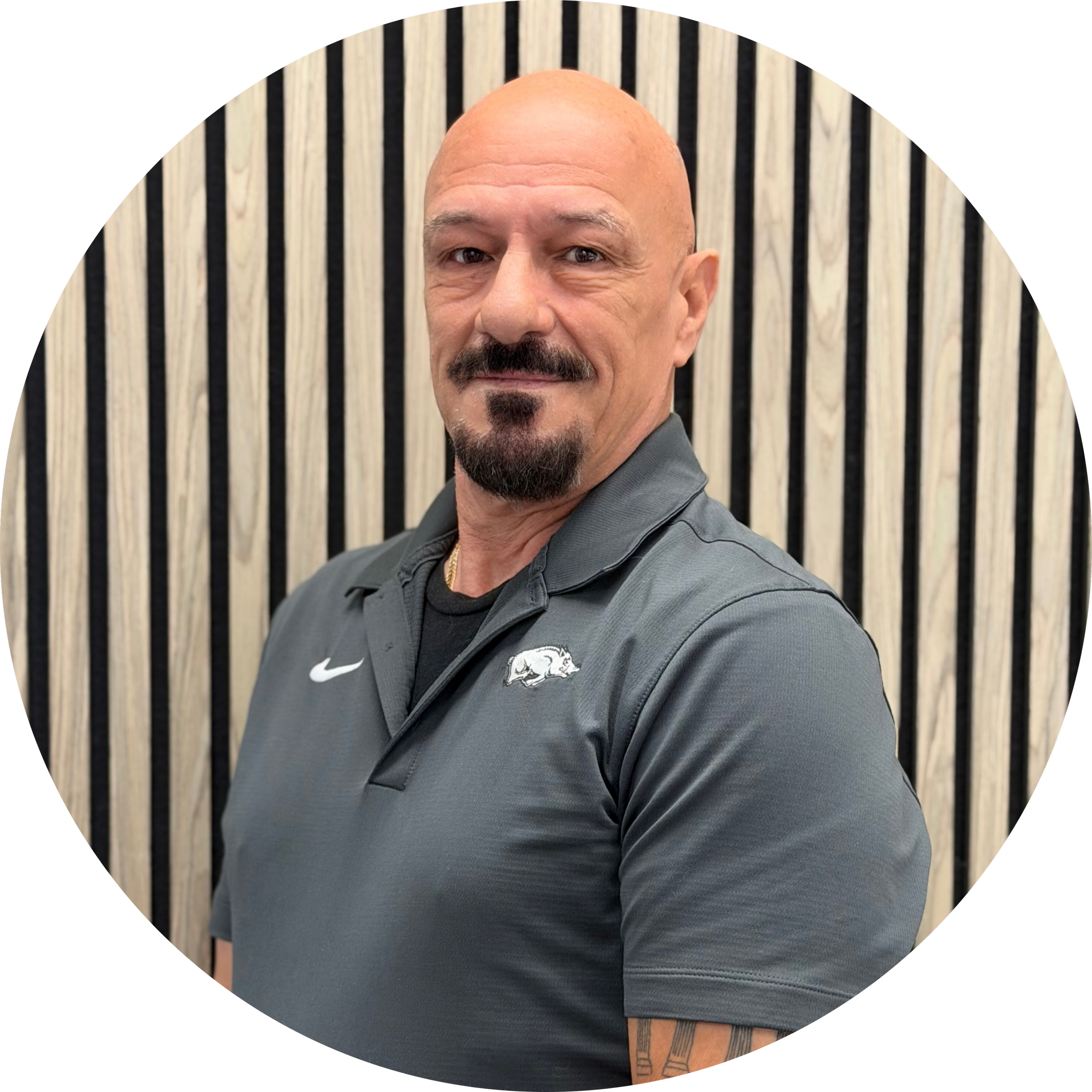 Man with a bald head, mustache, and goatee, wearing a gray shirt with a logo, standing in front of a background with vertical black and beige stripes.
