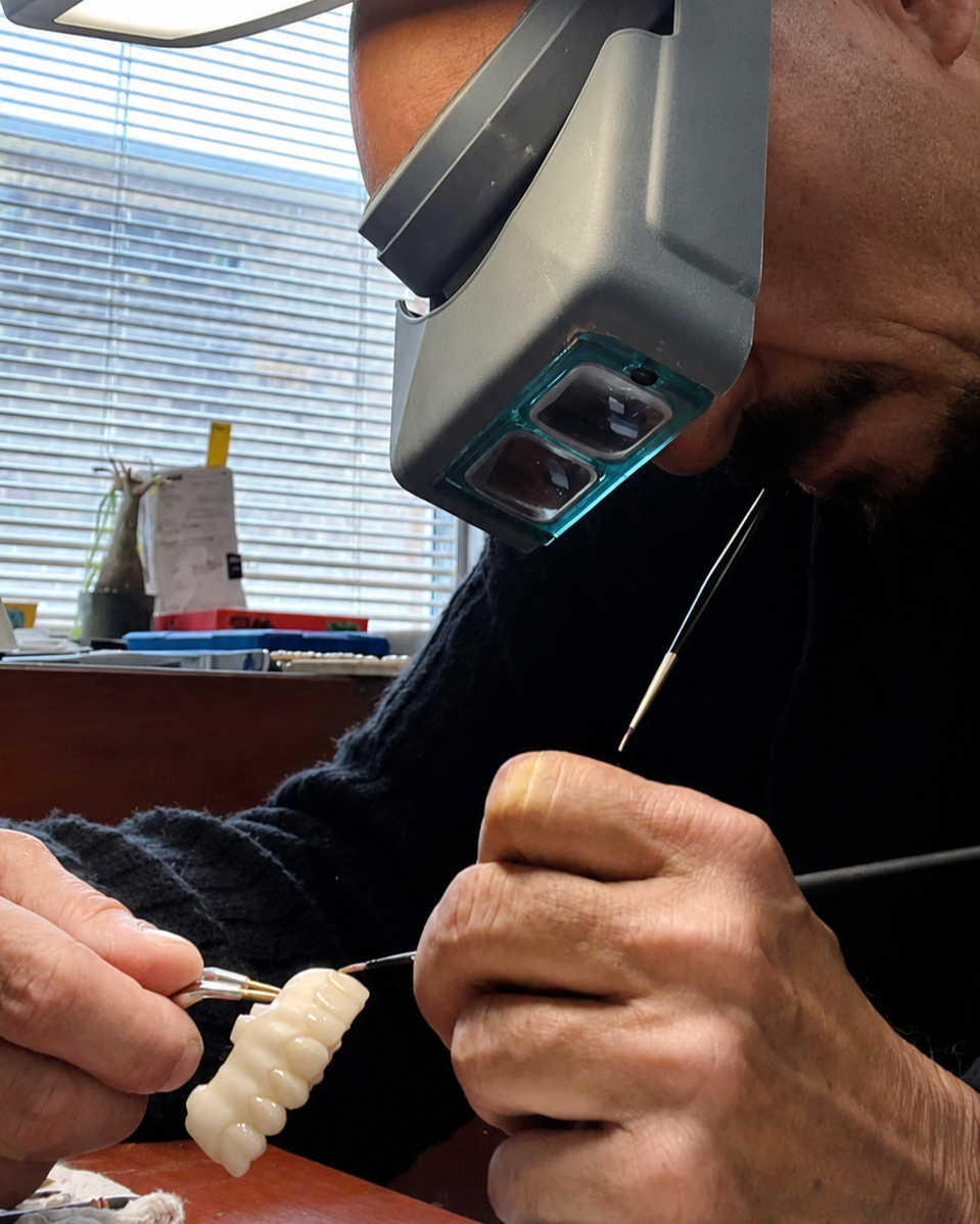A man working on a dental prosthetic, focusing intensely through magnifying glasses with lights, using fine tools in a dental lab or workshop.