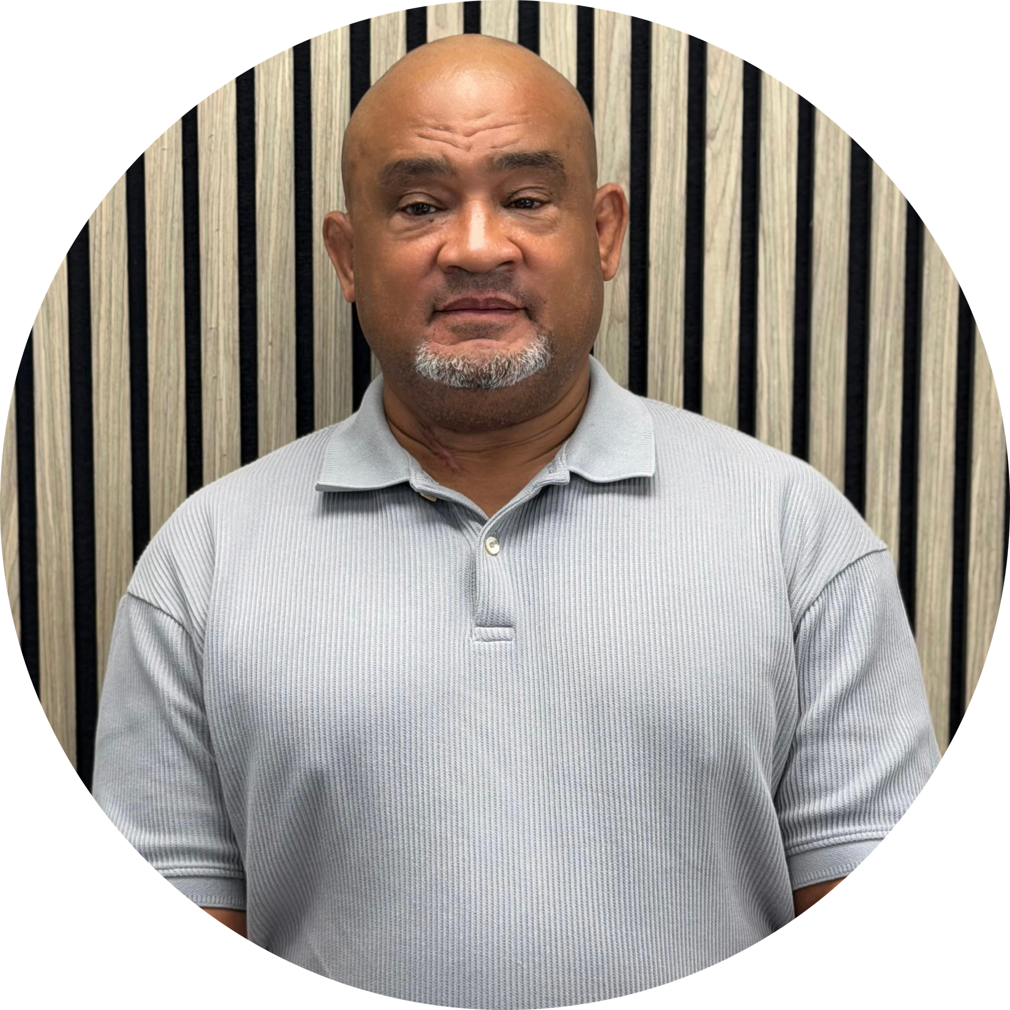 A middle-aged man with a shaved head and goatee, wearing a light gray polo shirt, standing against a background of black and beige vertical striped wooden panels.