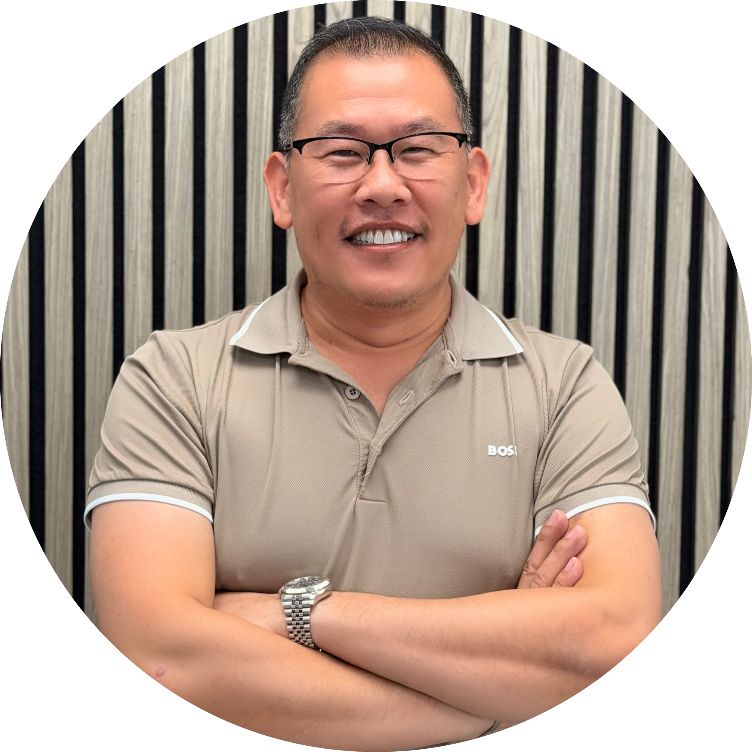 A smiling man with short dark hair, glasses, and a beige polo shirt with a small logo, standing with crossed arms against a vertical striped background.