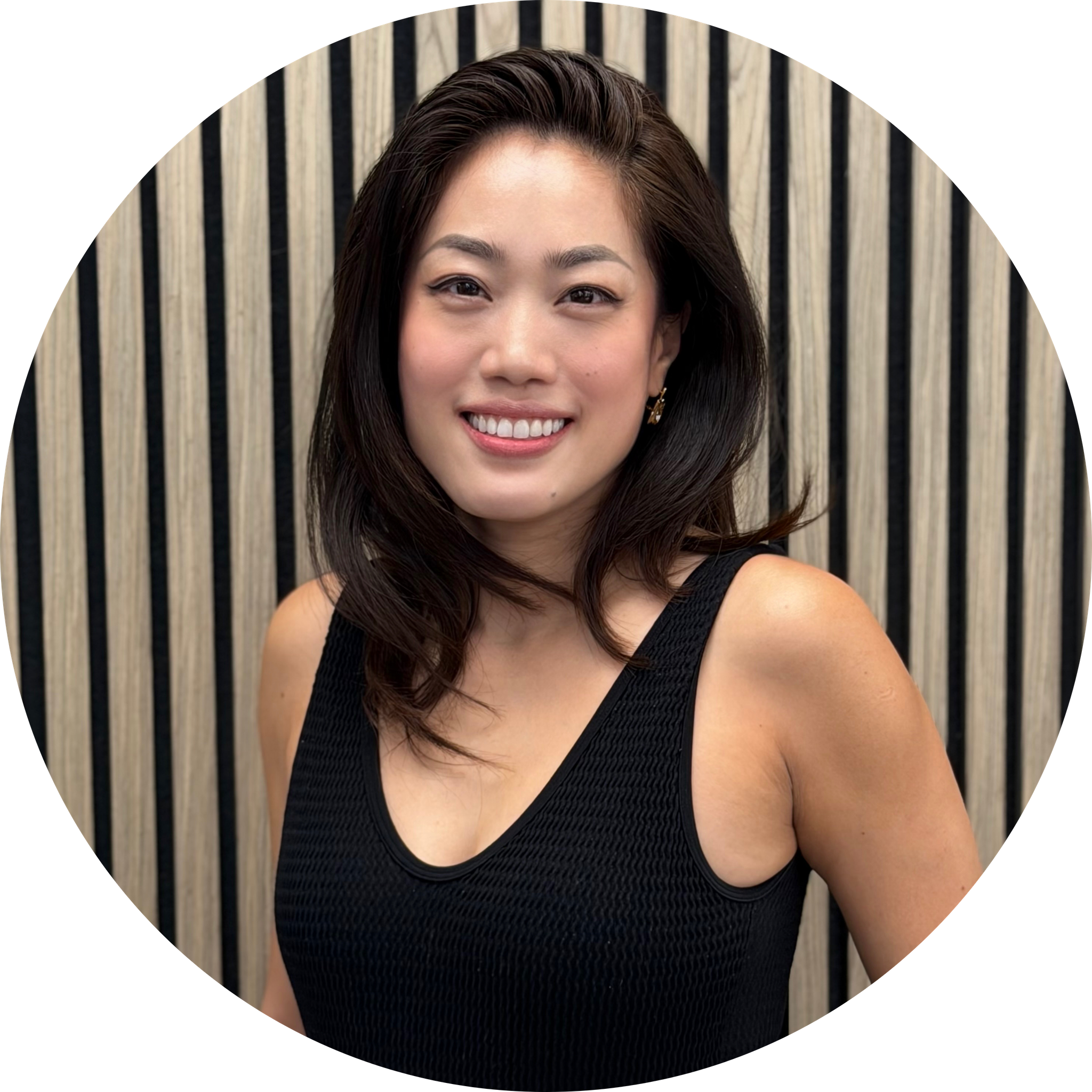A woman with dark brown hair, bright smile, wearing small gold earrings, black sleeveless top, standing in front of a vertical wood panel background.