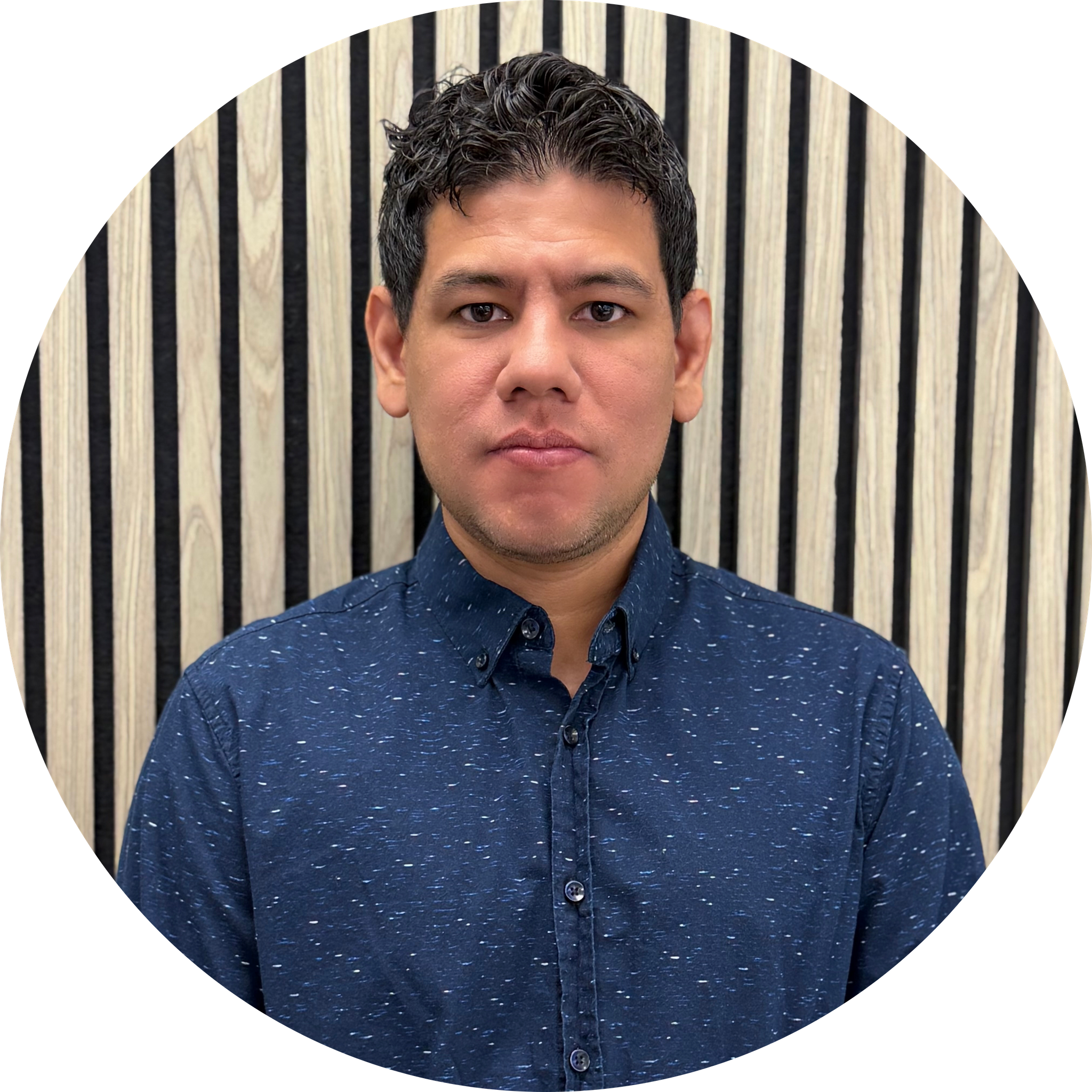 Close-up of a young man's face with short, curly dark hair, wearing a dark blue shirt with a subtle pattern, against a background of vertical black and natural wood-colored stripes.