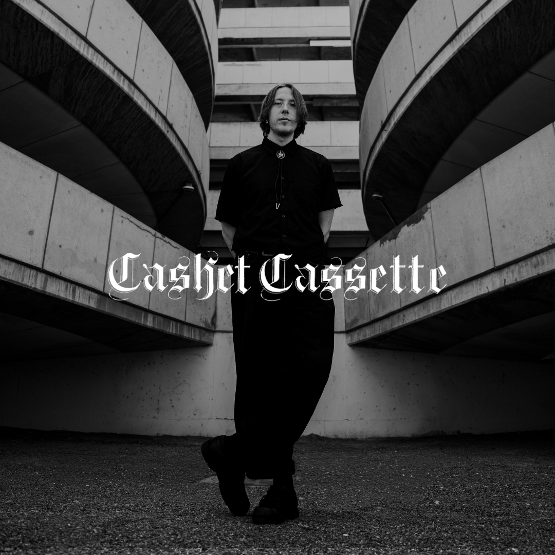 A man with shoulder-length hair standing between two curved modern concrete and steel structures in black and white, with gothic-style text "Cashot Cassette" overlayed.