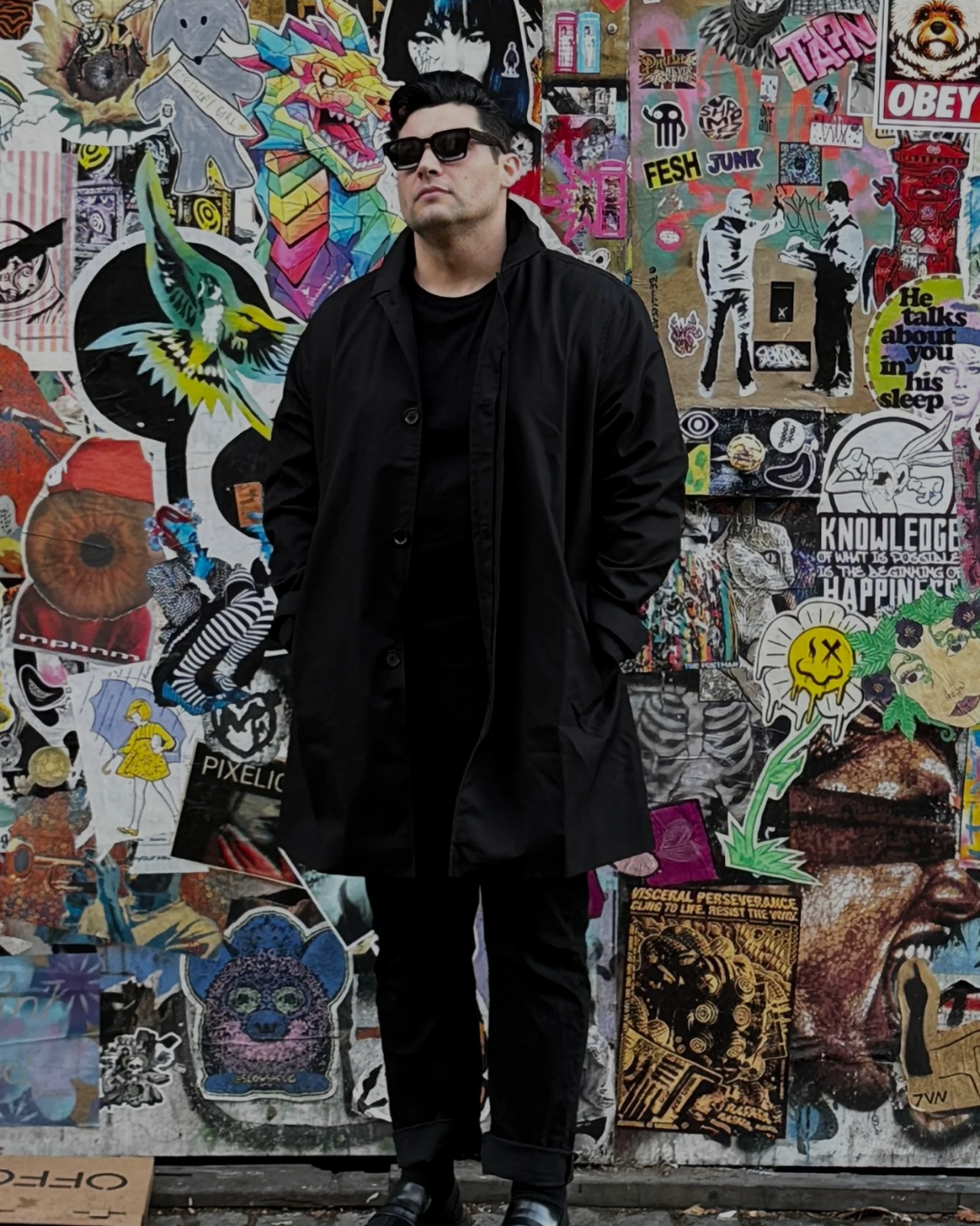A man dressed in all black with sunglasses stands in front of a vibrant wall covered in various colorful posters, stickers, and artwork.