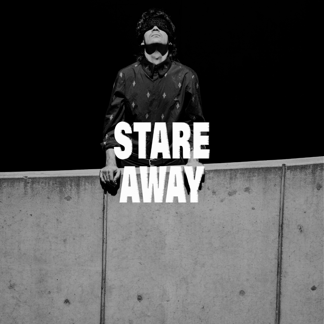 A person standing behind a concrete wall, wearing a patterned shirt and a band over their eyes, with the words "STARE AWAY" in bold white letters across the front.