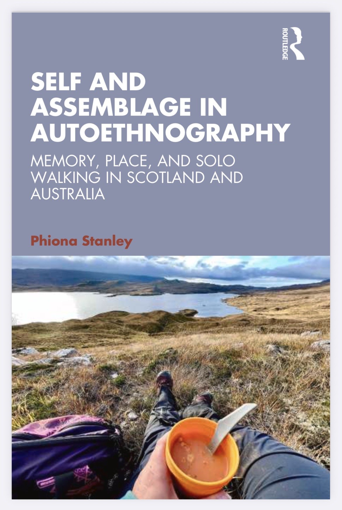 Book cover titled "Self and Assemblage in Autethnography" by Phiona Stanley, showing a landscape with a lake, mountains, and a person sitting on grassy terrain eating from an orange cup.