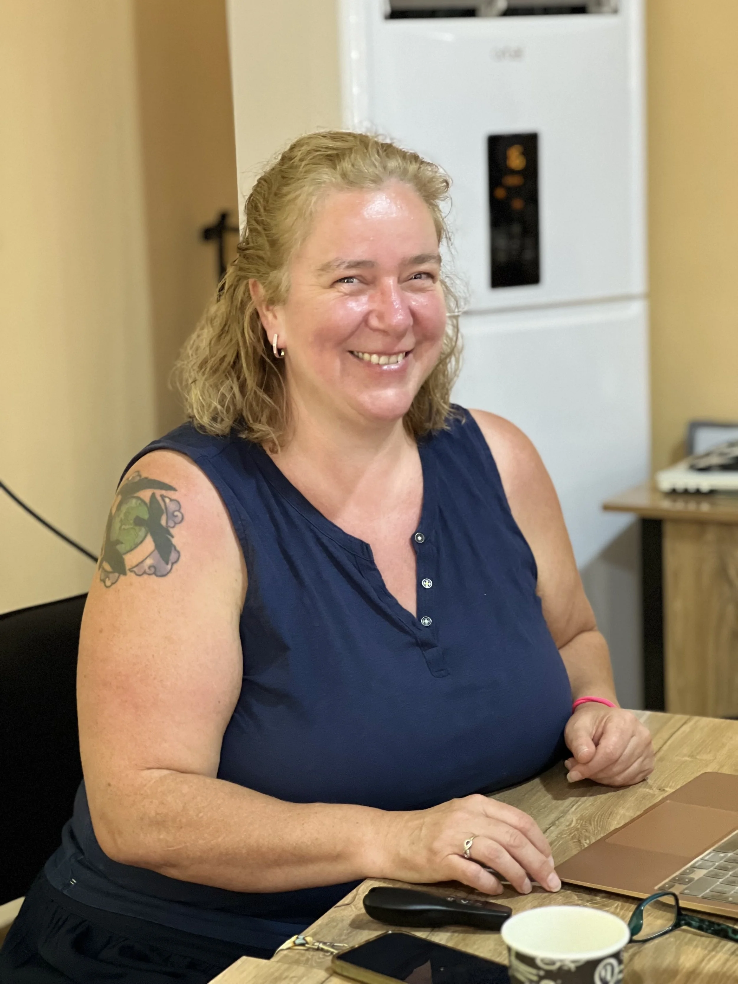 A smiling woman, Dr Phiona Stanley, wearing a blue sleeveless top, sitting at a wooden desk in a classroom with a laptop, a smartphone, a mug, and glasses in front of her. She has a colorful tattoo on her right arm.