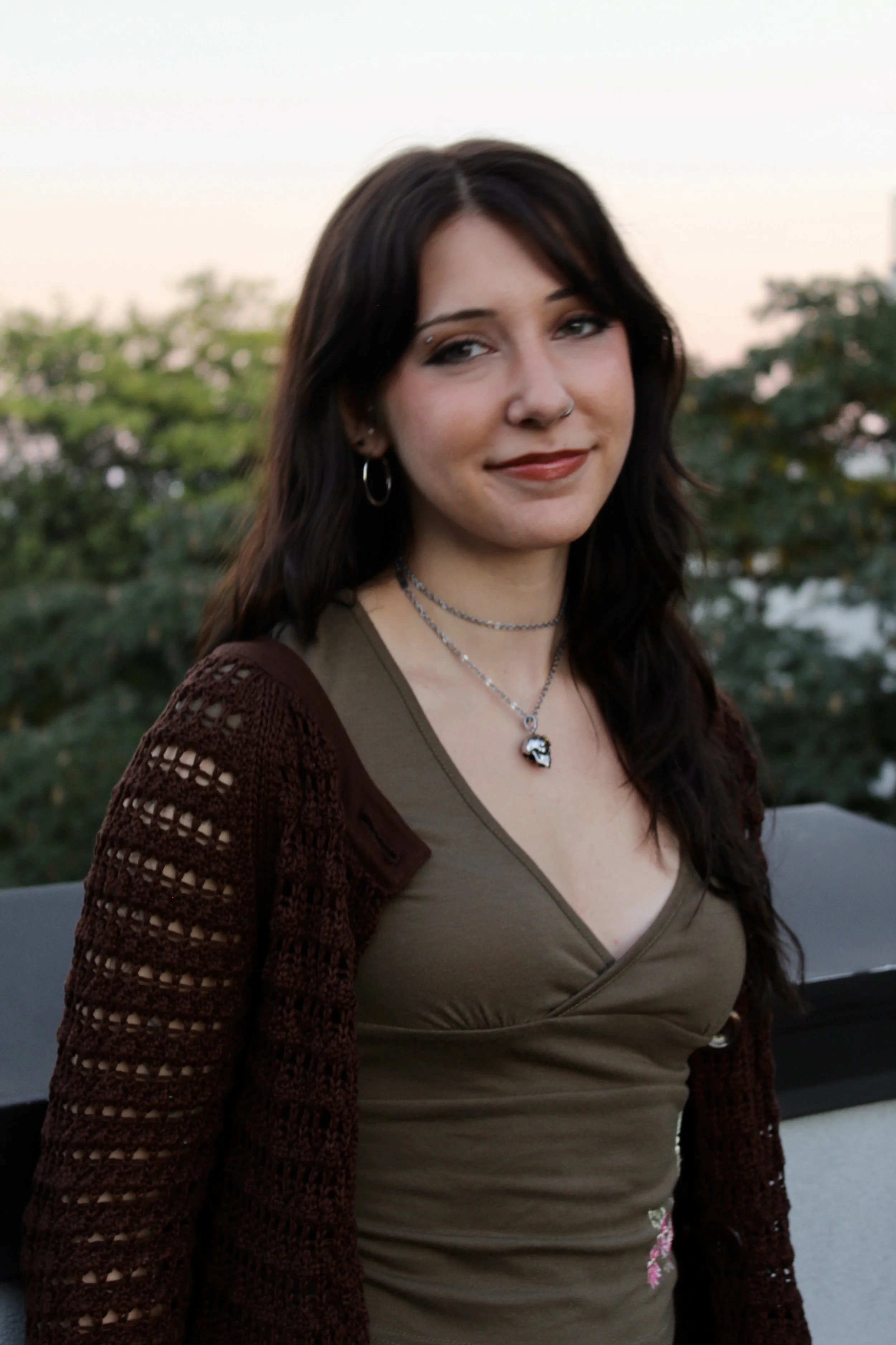A young woman with dark brown hair and multiple piercings, wearing a green top and brown crochet cardigan, standing outdoors during sunset with greenery in the background.