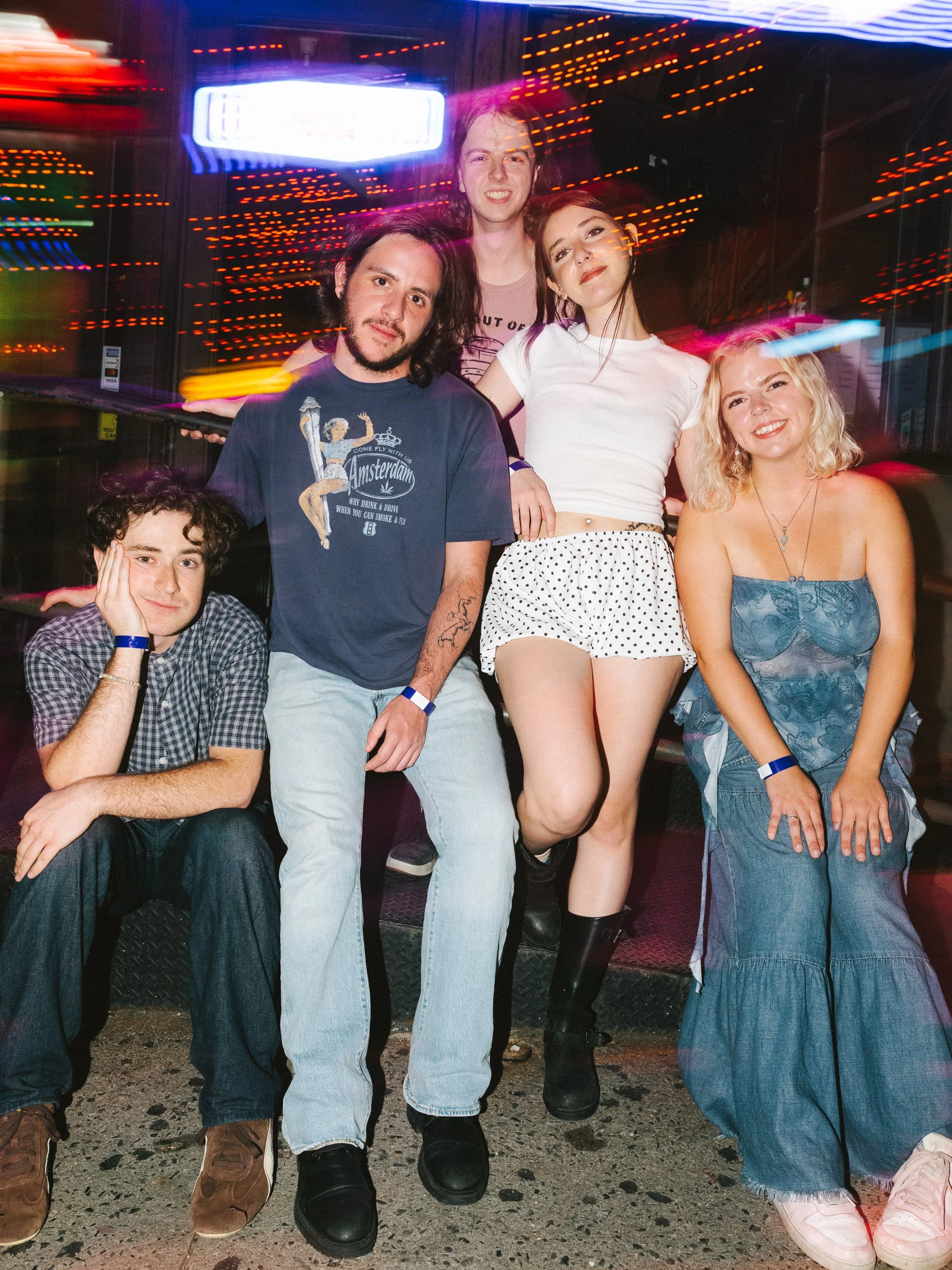 Group of five young adults posing together at night in front of neon lights, some with colorful light streaks across the image.