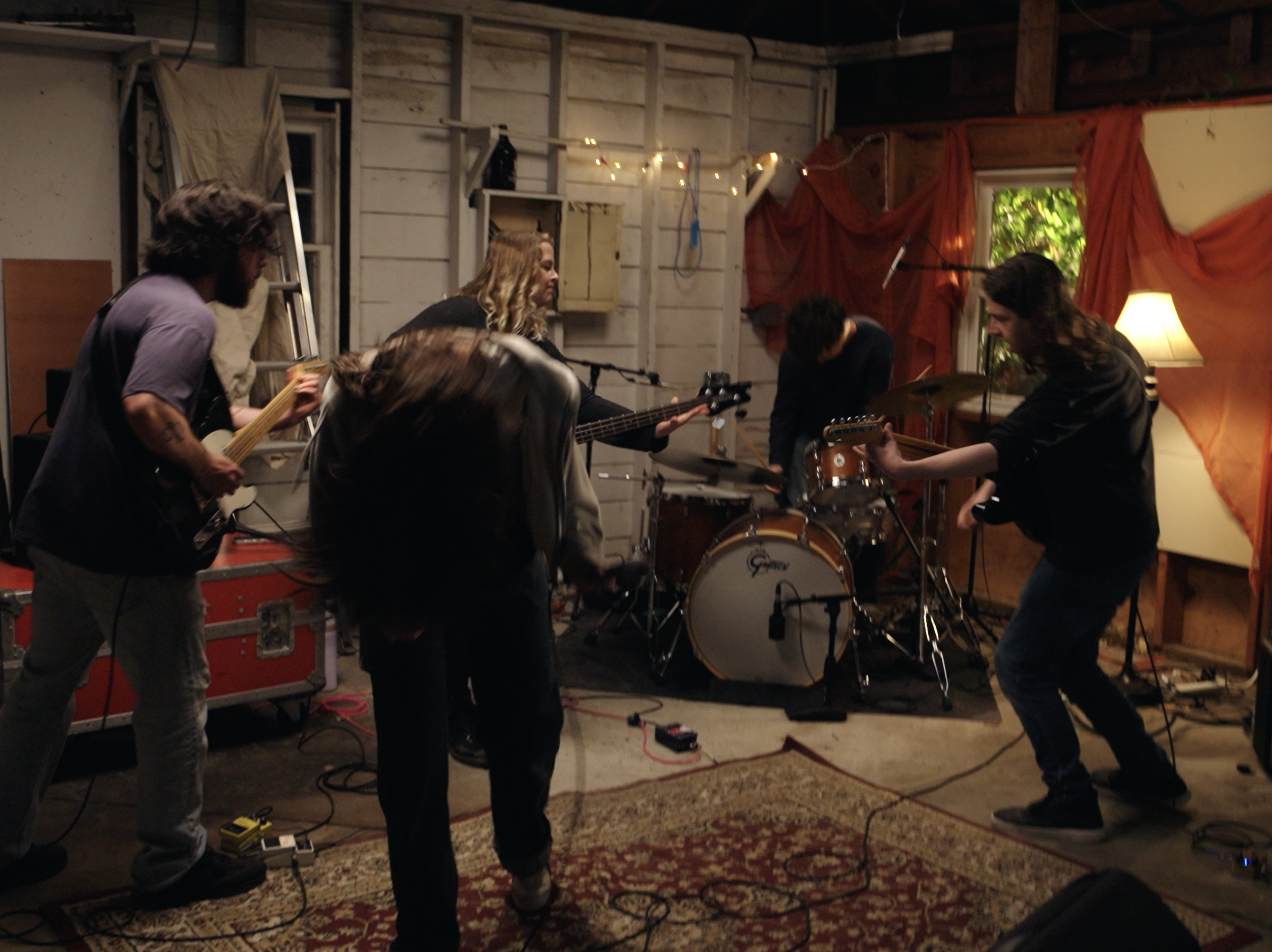 A group of five people playing musical instruments in a rustic indoor setting. Two individuals are playing guitars, one is playing a keyboard, and there is a drum set. The room is decorated with string lights, a lamp, and orange curtains, with a window showing greenery outside.