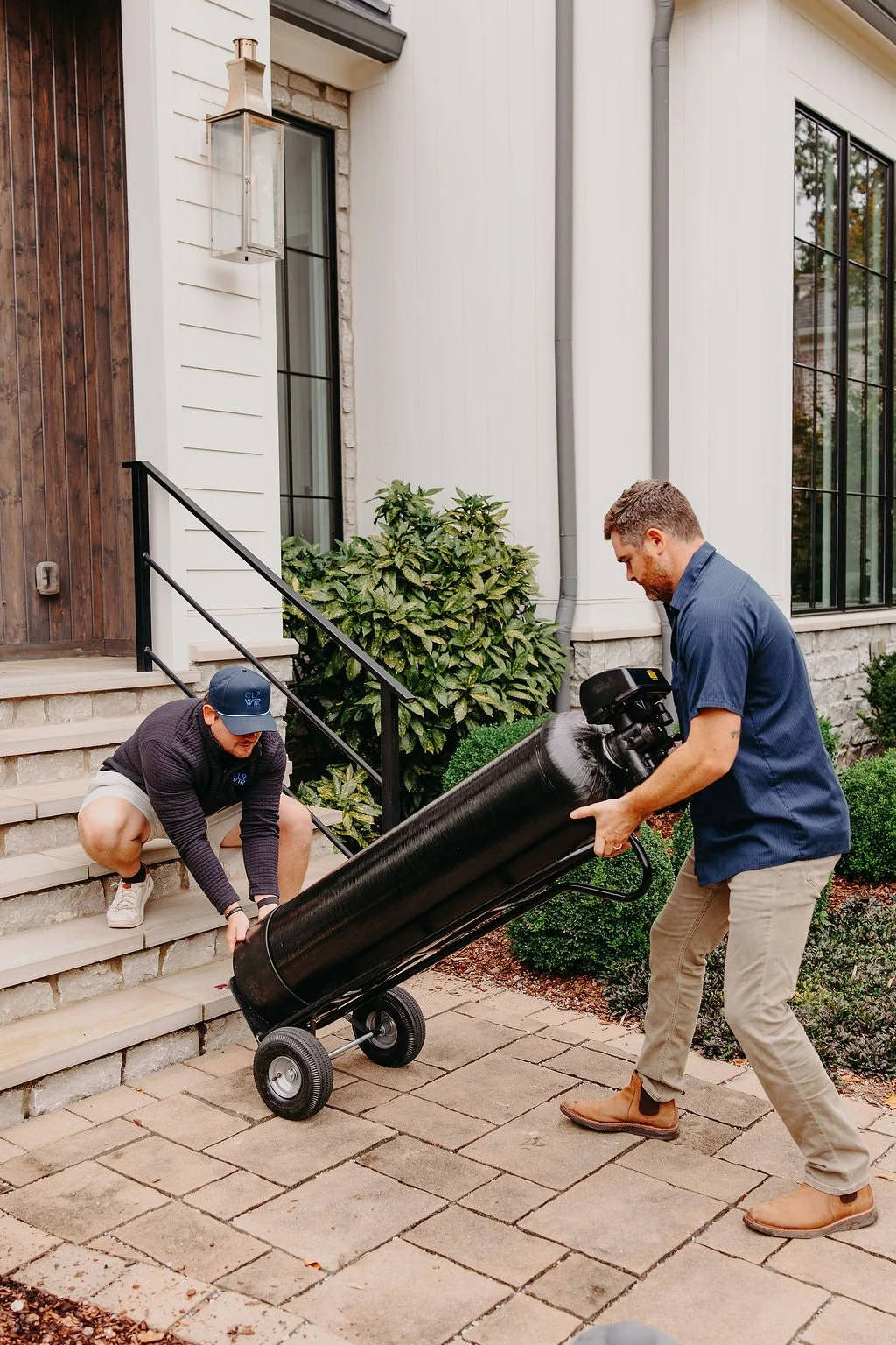 Jon Smith and Jared of CLR WTR Solutions carrying a water filtration tank up porch stairs to a home for installation