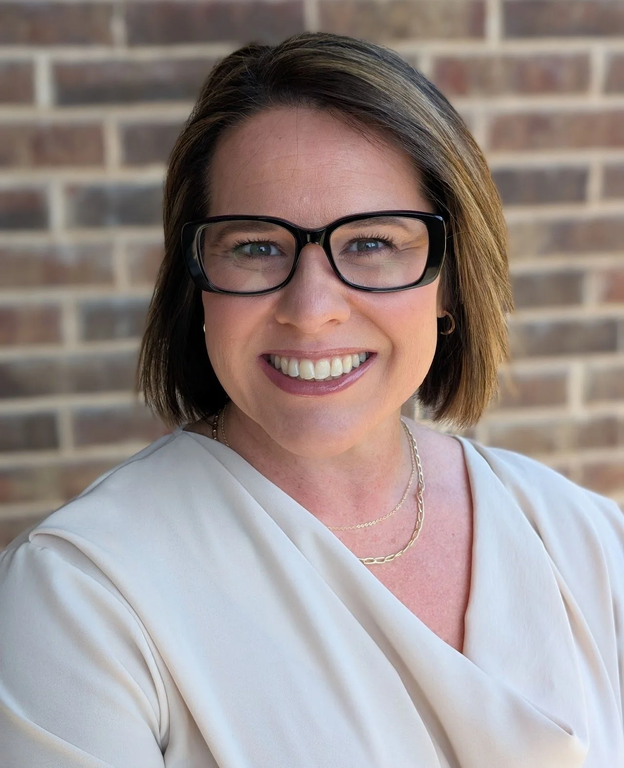 A woman with short brown hair, glasses, and a white blouse, smiling in front of a brick wall.