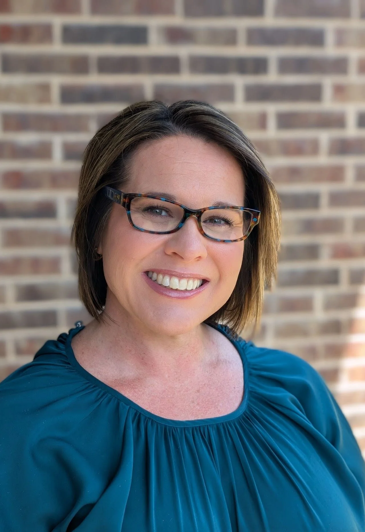 A woman smiling in front of a brick wall, wearing glasses and a teal blouse.
