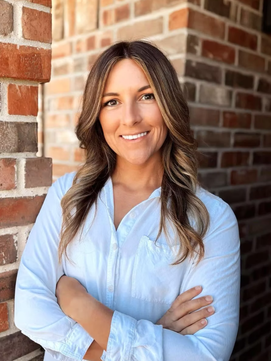 Portrait of a woman with wavy brown hair and a light blue button-up shirt, standing confidently with arms crossed in front of a brick wall exterior.