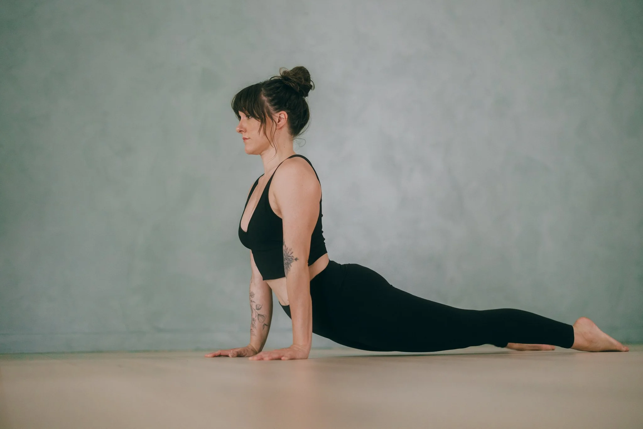 A woman in black activewear performing a yoga pose on a wooden floor against a plain gray wall, in a yoga studio.
