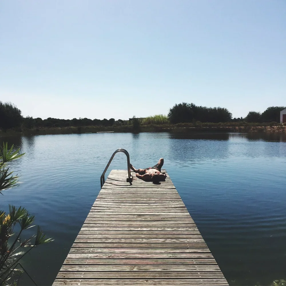 Person lying on a dock by a calm lake, with trees and clear sky in the background.