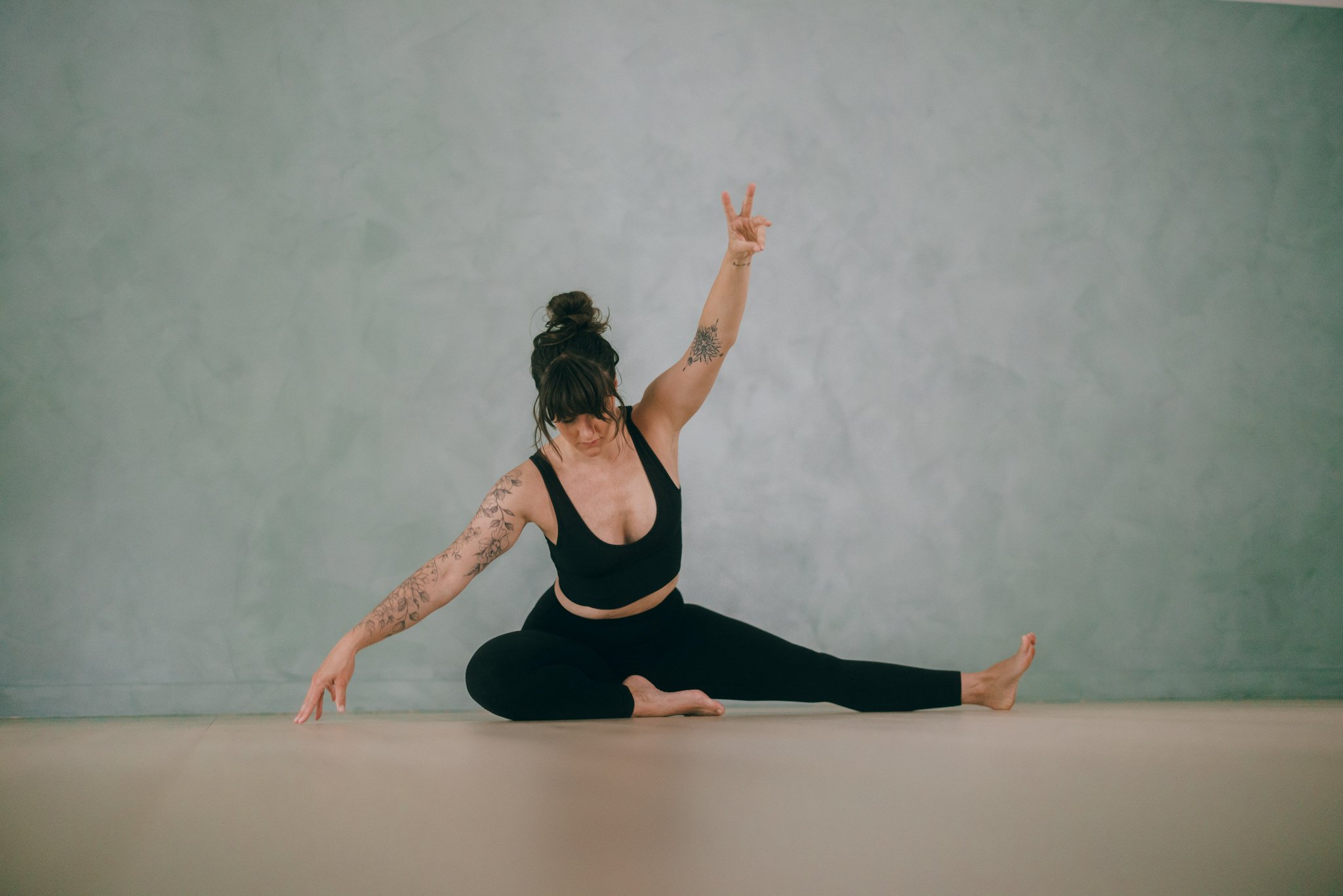 A woman practicing yoga in a yoga studio, seated in a cross-legged pose with her right arm raised and making a peace sign, wearing a black sports bra and leggings, with tattooed arms and dark hair tied up in a bun, against a plain light-colored wall.