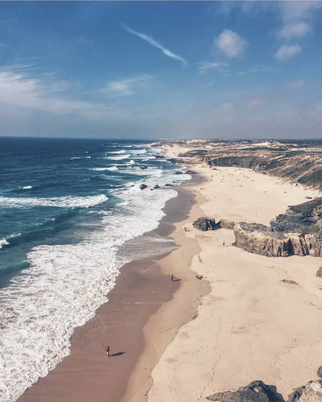 A scenic view of a beach with sandy shore and cliffs, waves crashing on the beach, and a blue sky with some clouds.